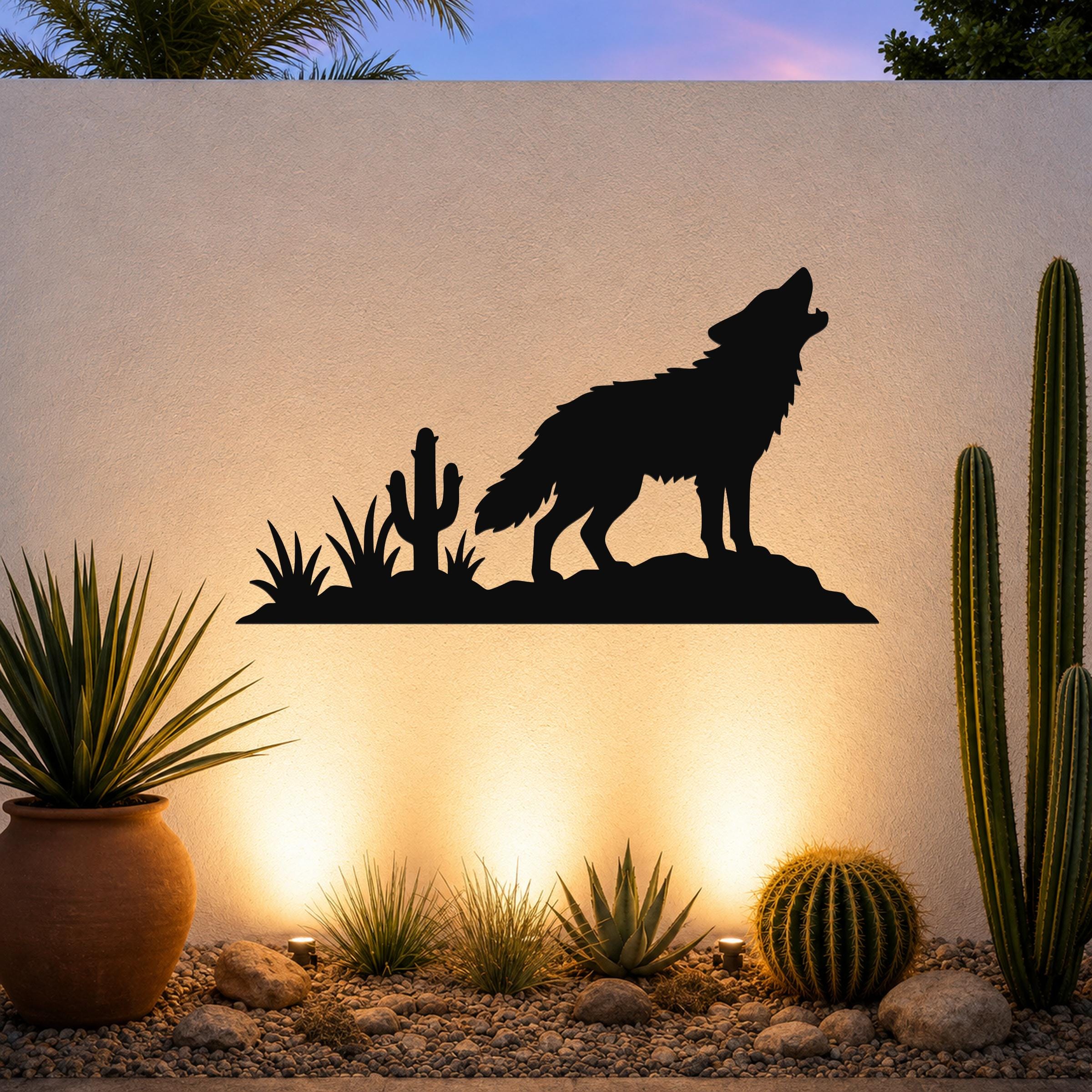 Black metal wall art silhouette of a coyote howling on a rock with cacti, mounted on an outdoor wall above a desert garden with warm uplighting.