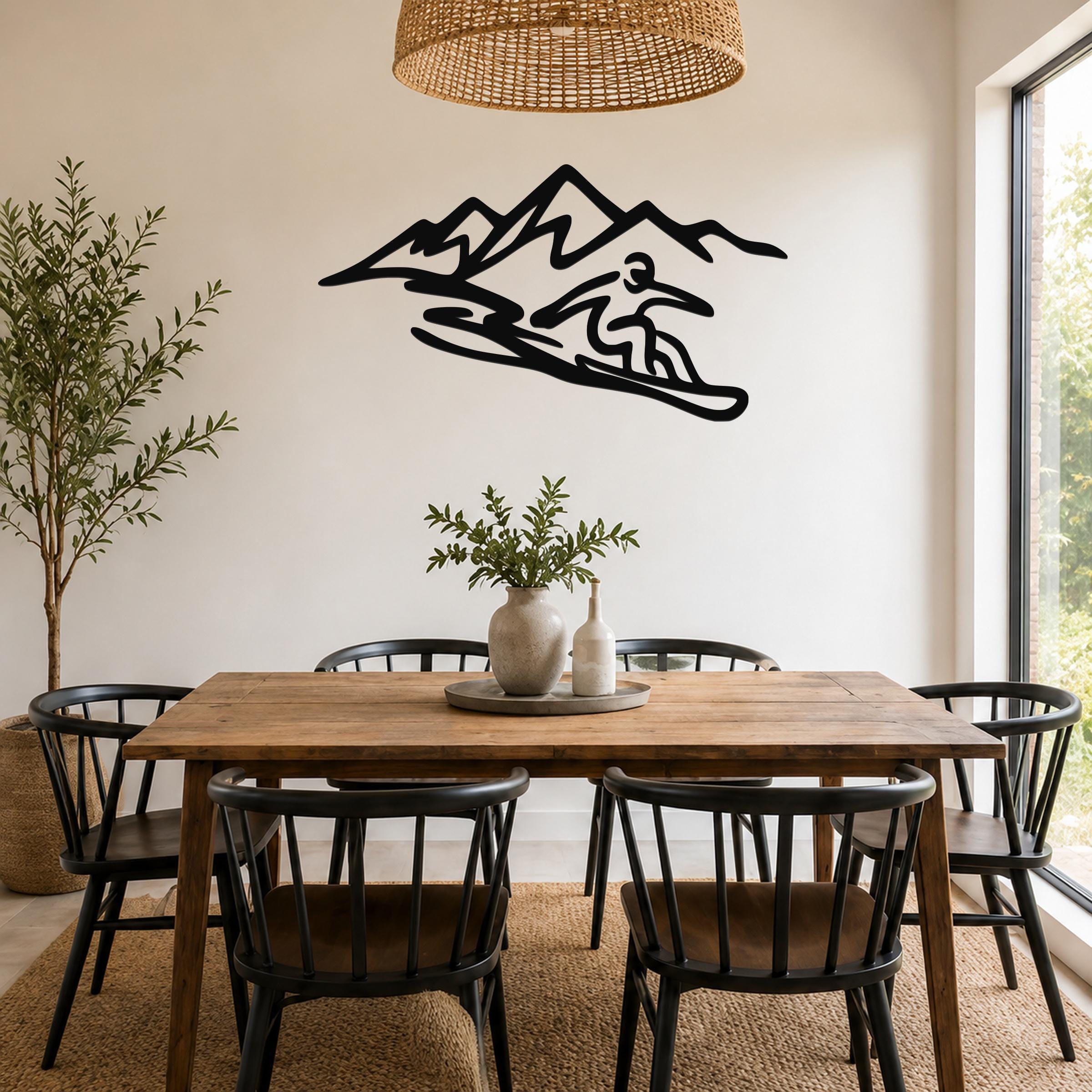 Black metal wall art silhouette of a snowboarder in motion against a mountain range, mounted on a white wall above a wooden dining table in a bright room.
