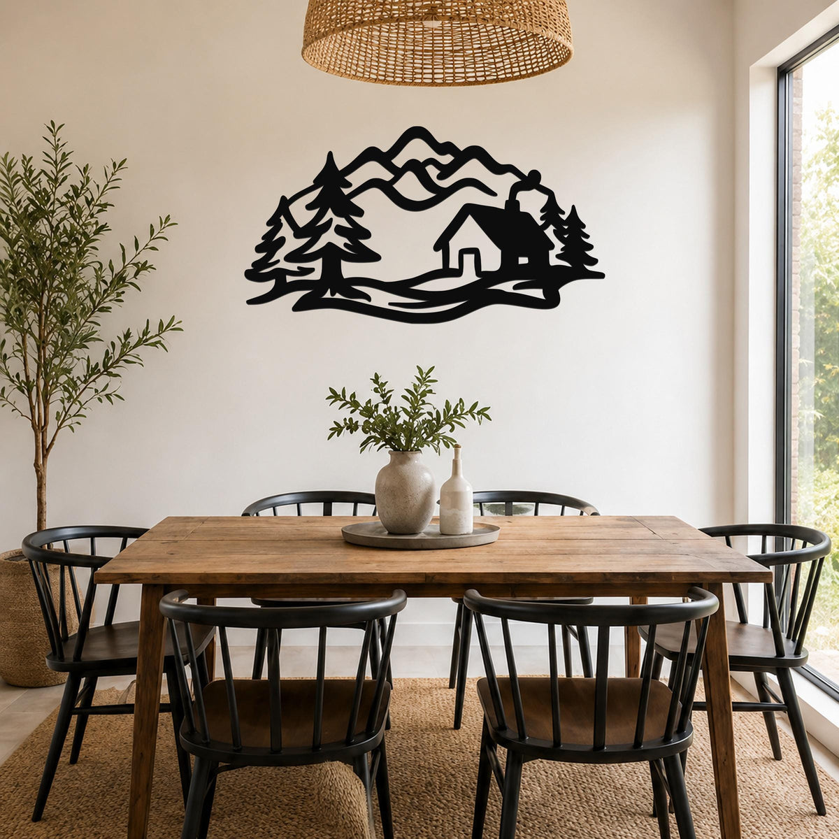 Black metal wall art silhouette of a mountain cabin surrounded by trees, mounted on a wall above a wooden dining table in a bright room.