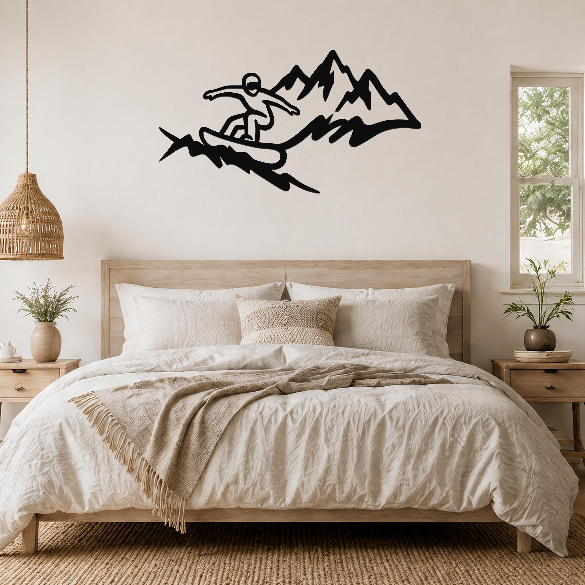 Black metal wall art featuring a snowboarder silhouette in motion above a mountain range, mounted on a bedroom wall above a wooden bed.