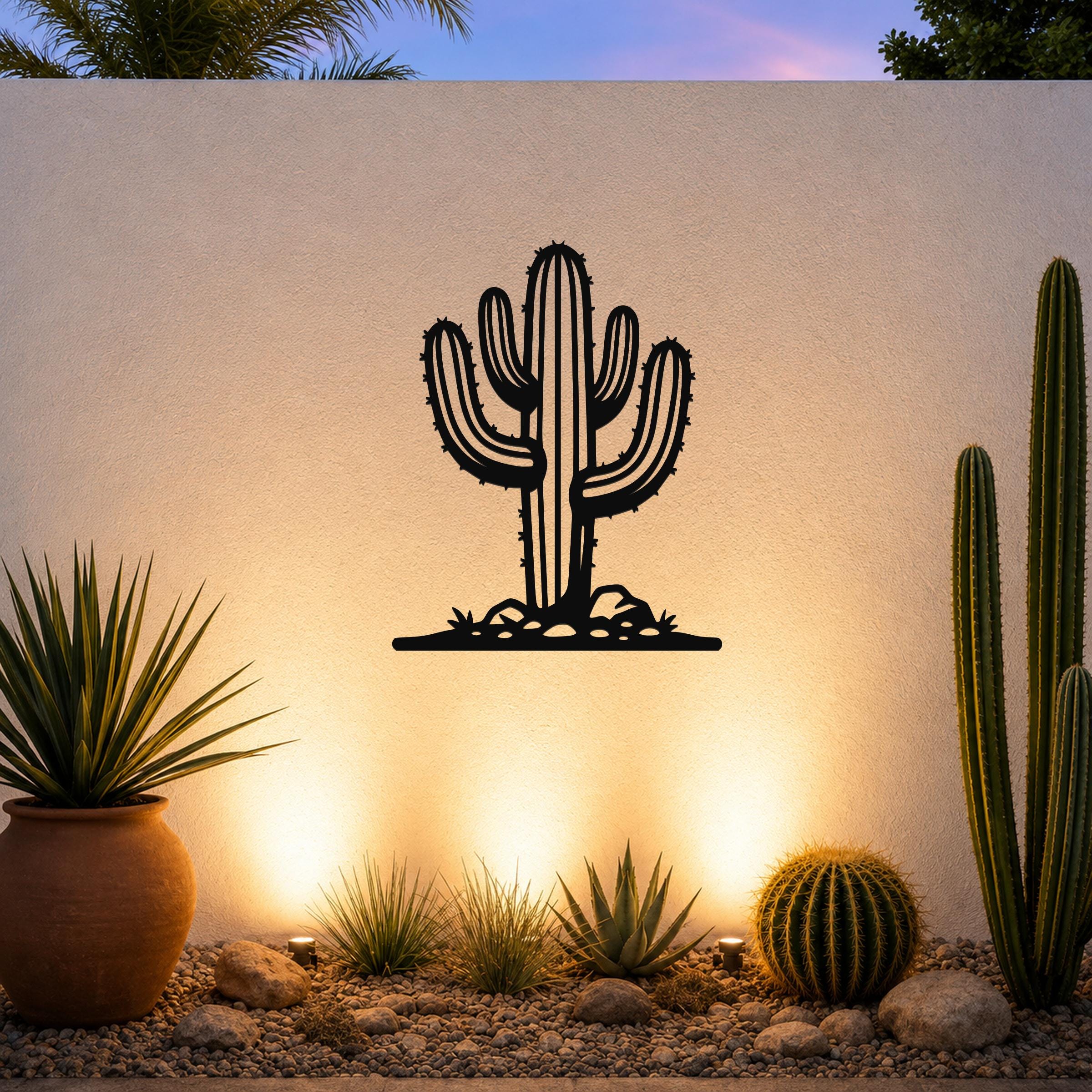 Black metal wall art silhouette of a saguaro cactus with branching arms and spines, mounted on an outdoor wall above a desert garden with potted plants and ground lights.