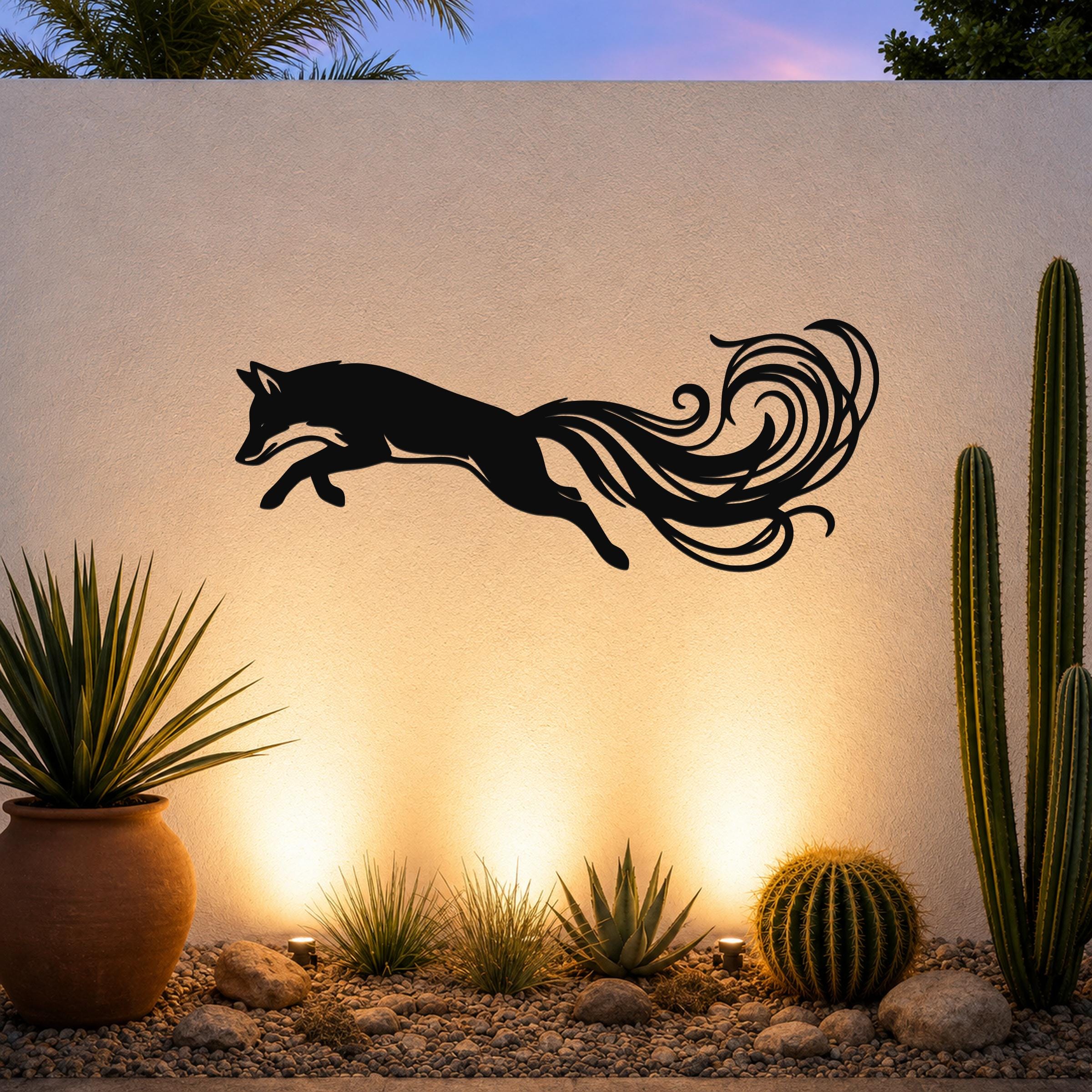 Black metal fox wall art with flowing tail cutouts, mounted on an outdoor wall above a desert garden with cacti and warm uplighting.