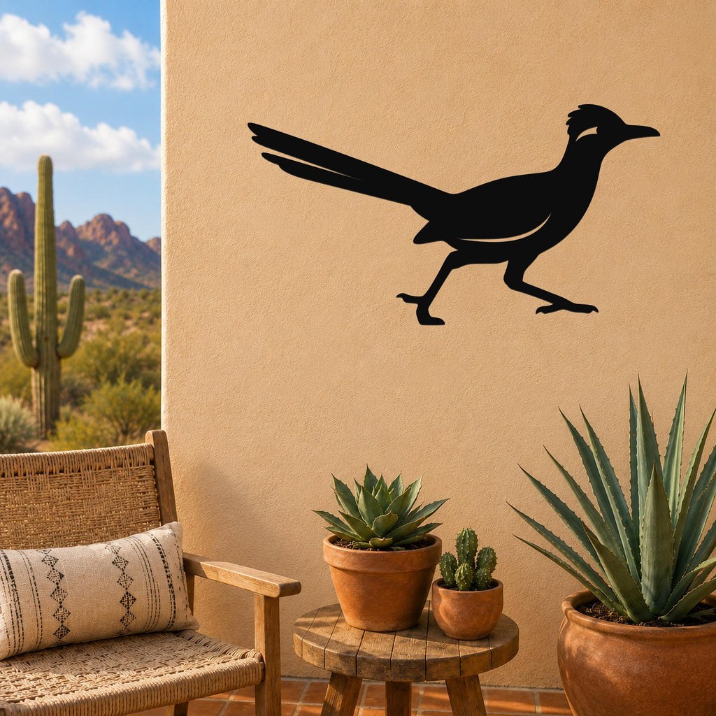 Black metal roadrunner wall sculpture with long tail and crest silhouette, mounted on a tan outdoor wall above a small wooden table with potted succulents.