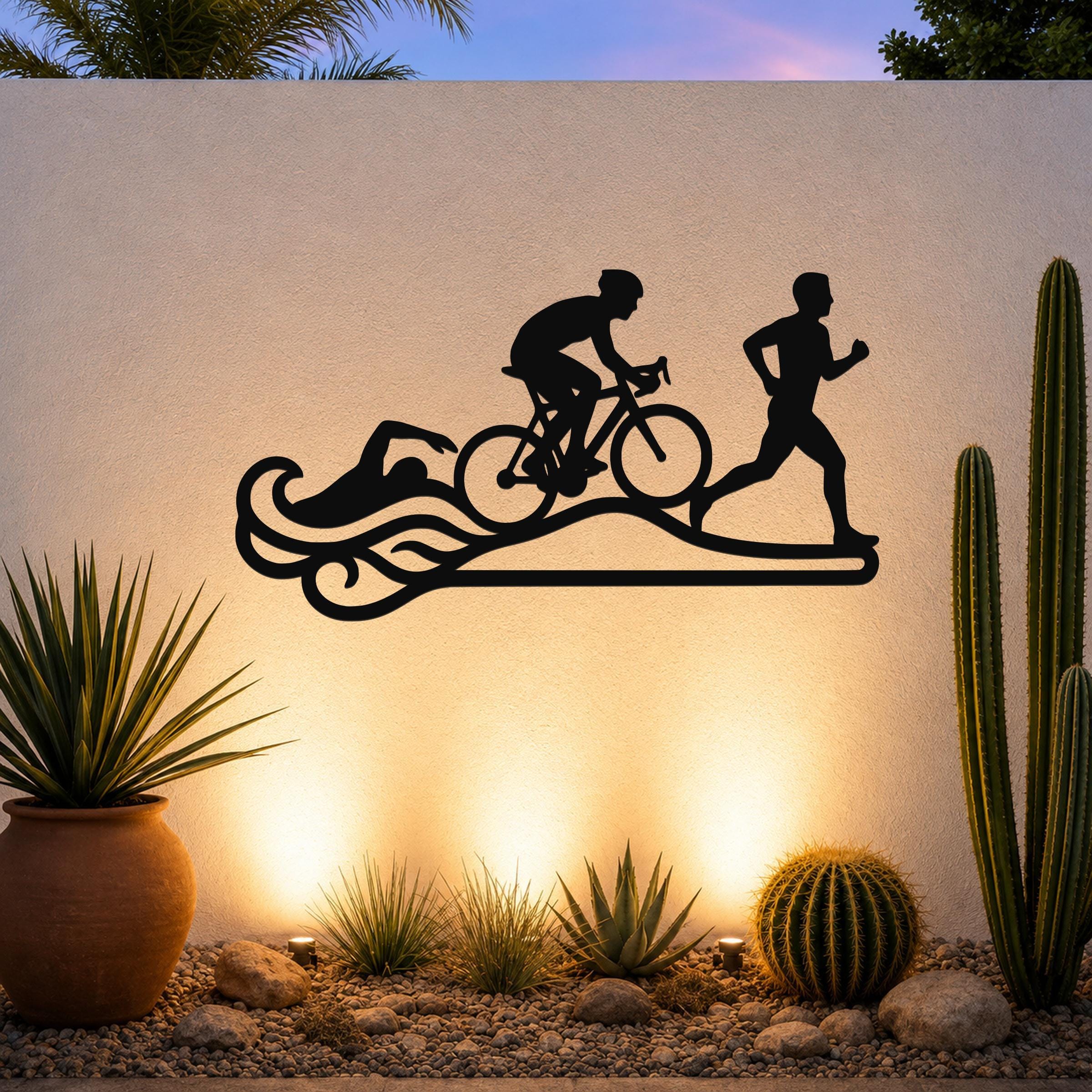 Black metal triathlon wall art with swim, bike, and run silhouettes and wave cutouts, mounted on an outdoor wall above a desert garden with cacti and warm uplighting.