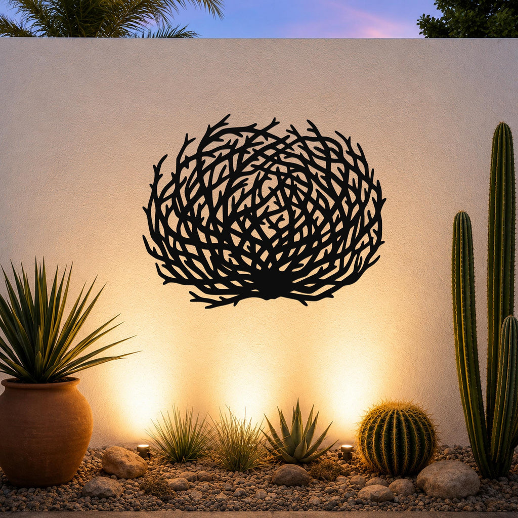 Black metal wall art with a circular, branch-like cutout design, mounted on an outdoor wall above a desert garden with cacti and warm uplighting.
