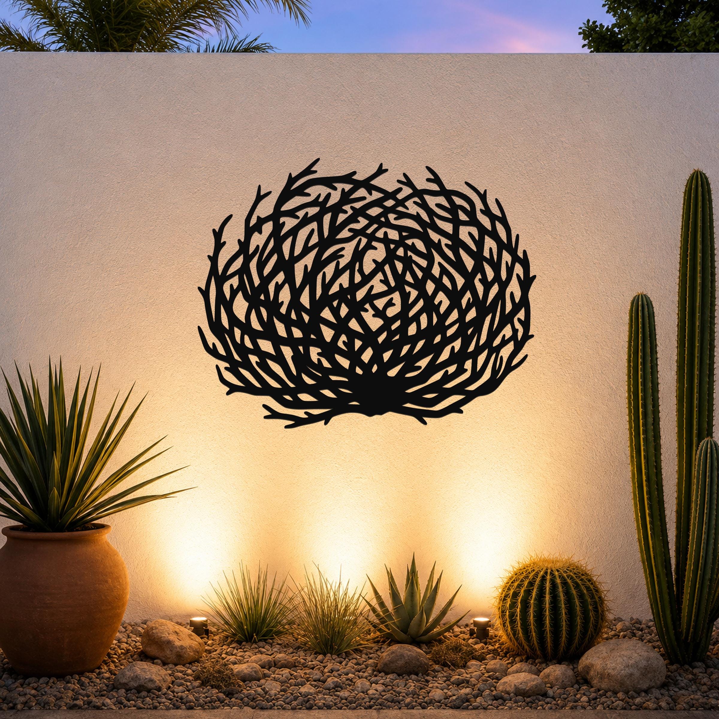Black metal wall art with a circular, branch-like cutout design, mounted on an outdoor wall above a desert garden with cacti and warm uplighting.