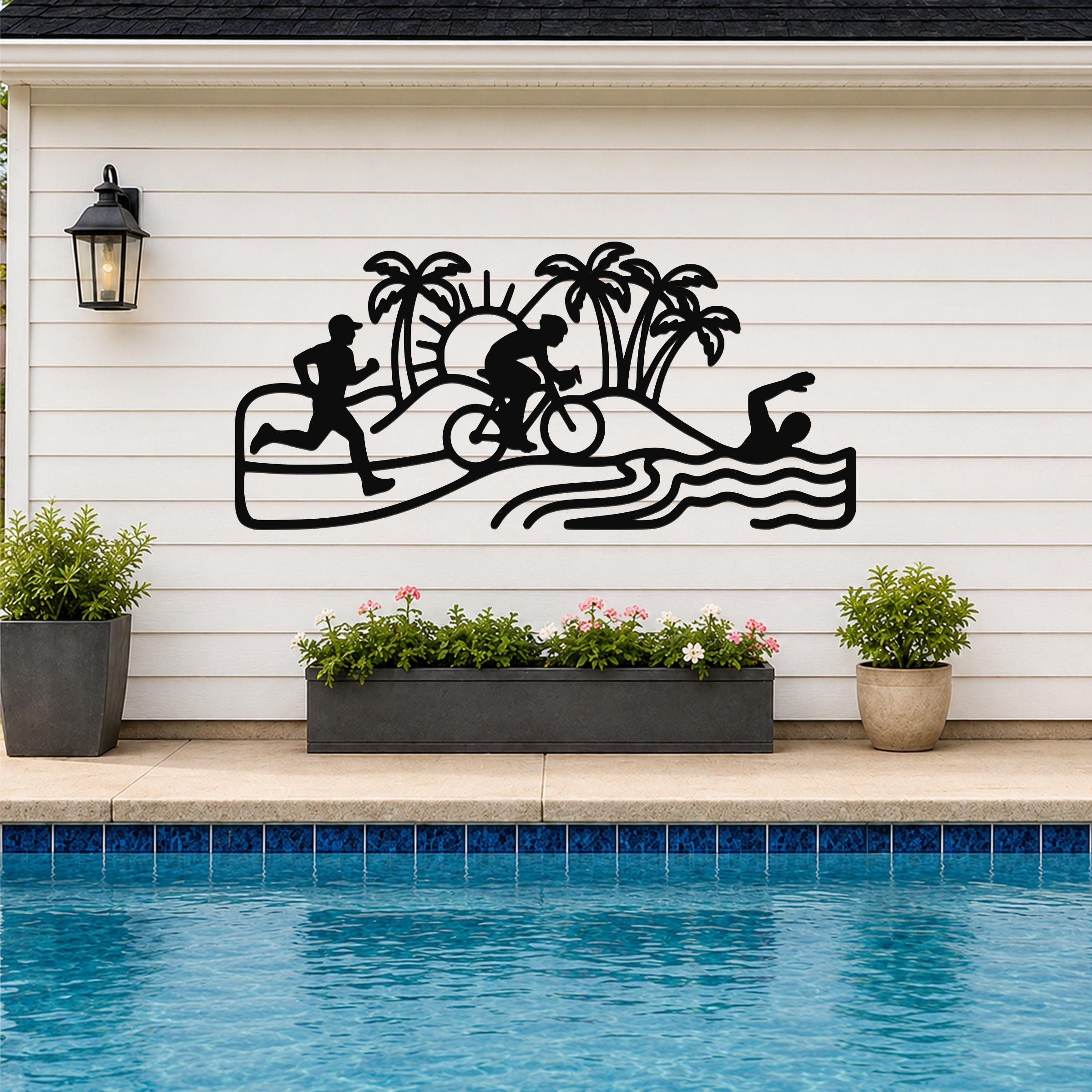 Black metal triathlon wall art with swim, bike, and run silhouettes, palm trees, and sun cutouts, mounted on a white siding wall above a pool.