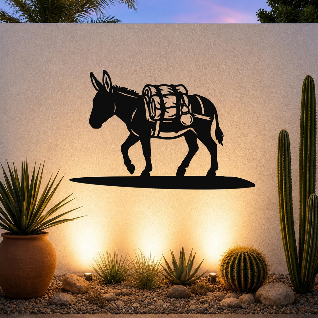 Black metal burro wall art with a backpack silhouette, mounted on an outdoor wall above a desert garden with cacti and warm uplighting.