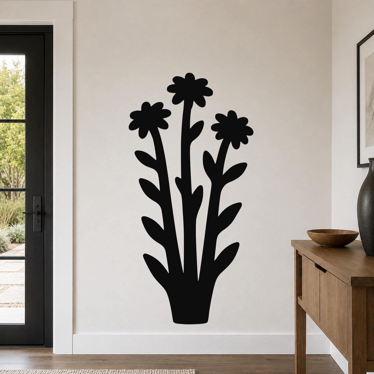 Black metal wall art silhouette of three wildflowers with layered petals and leaves, mounted on a light-colored wall in a living room near a door and console table.