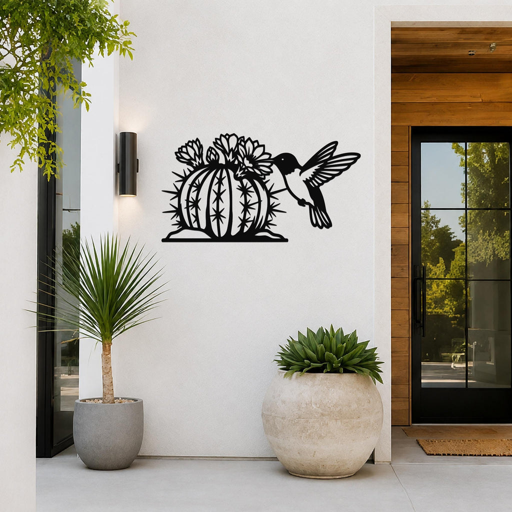 Black metal barrel cactus wall art with flowering blooms and a hummingbird cutout, mounted on a white exterior wall beside a glass door and potted plants.