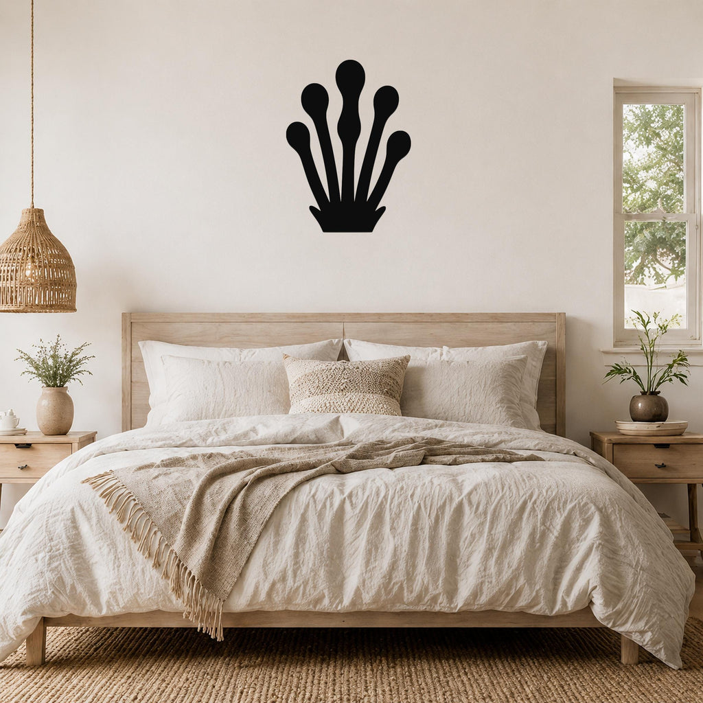 Black metal wall art in a silhouette of a kelp or seaweed frond with five rounded blades, mounted above a wooden bed in a neutral bedroom.