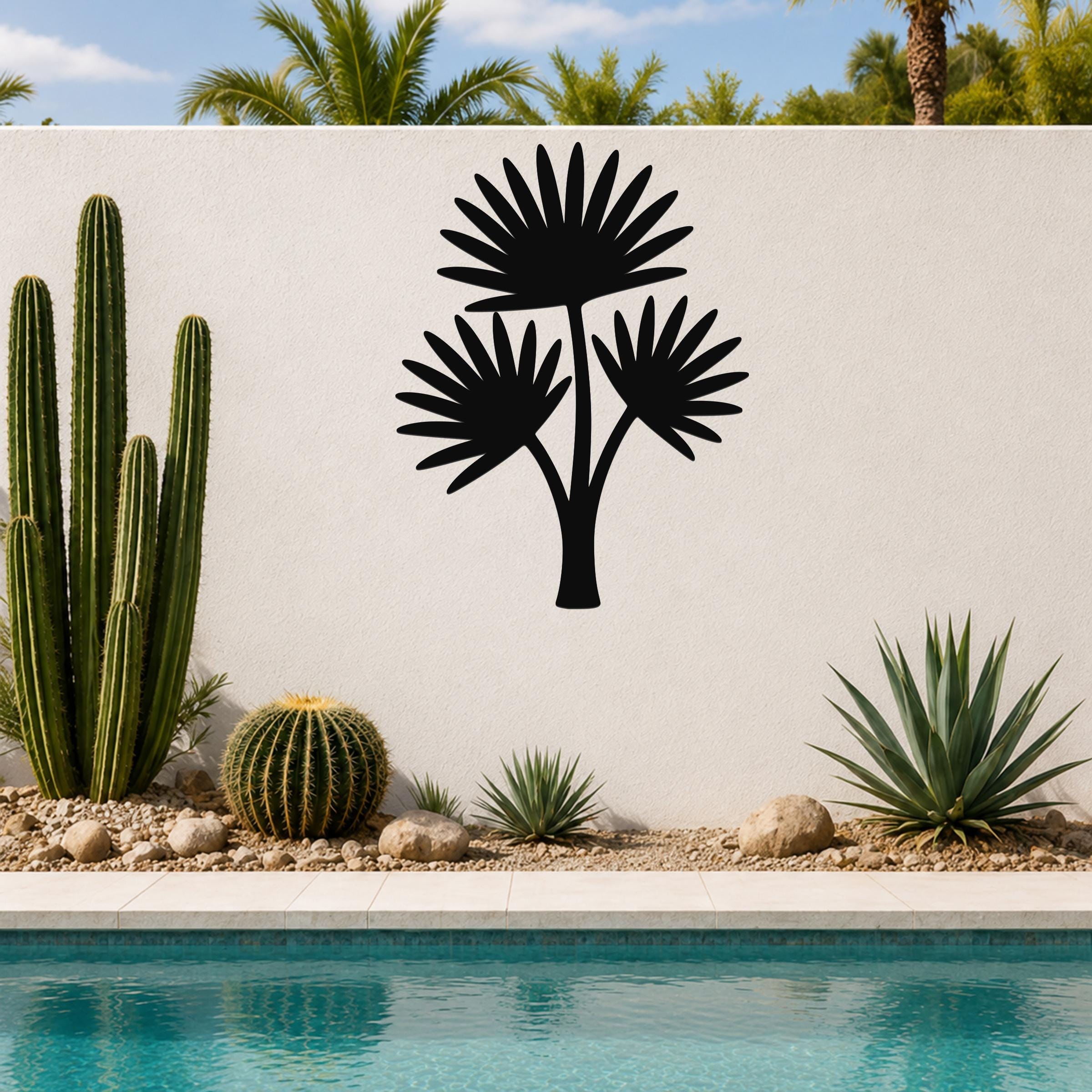 Black metal cutout wall art of a fan palm tree with three fronds, mounted on a white outdoor wall above a poolside garden with cacti and succulents.