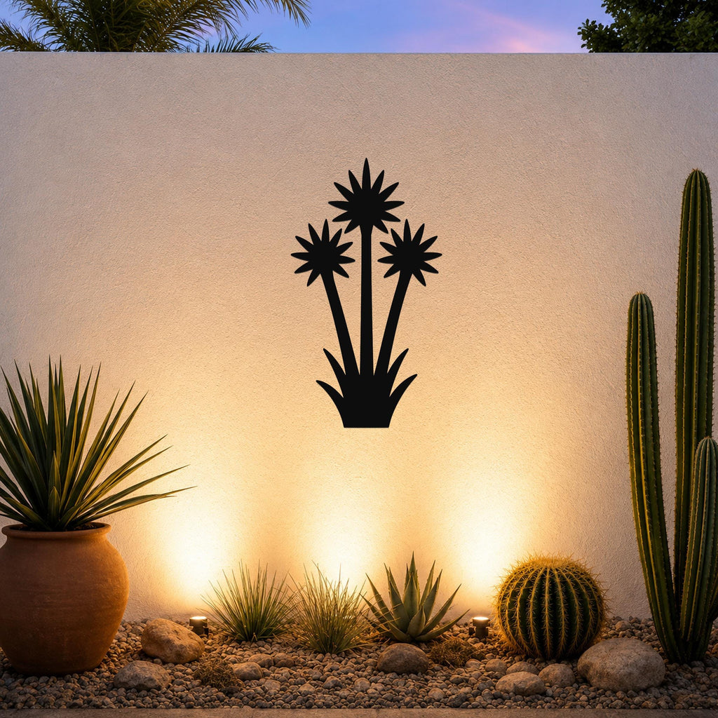 Black metal wall art cutout of a blooming yucca plant with three star-shaped flowers, mounted on a textured outdoor wall above a desert garden with potted succulents and ground lights.