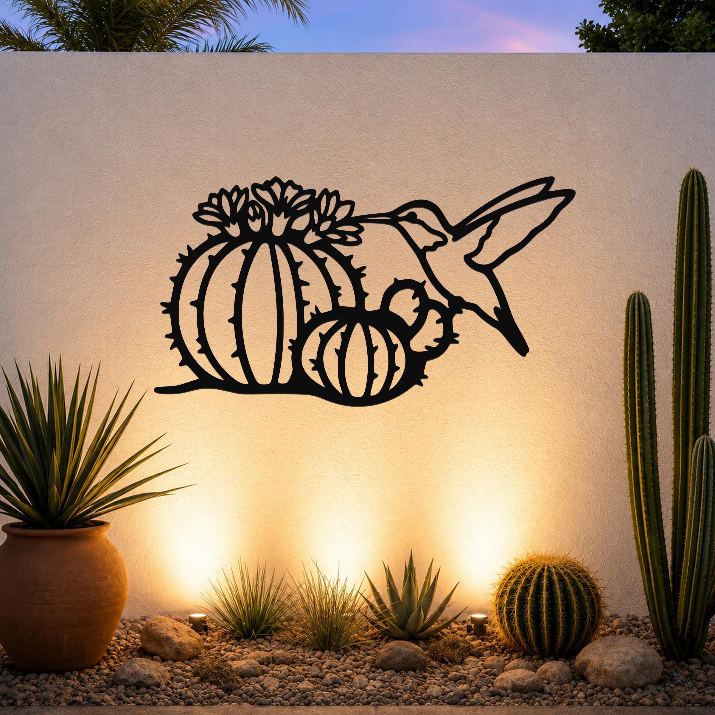 Black metal wall art featuring a hummingbird and barrel cactus with flower cutouts, mounted on an outdoor wall above a desert garden with potted plants and ground lights.