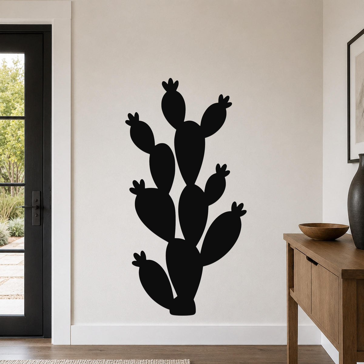 Black metal prickly pear cactus wall art with multiple segmented pads and small flower cutouts, mounted on a light-colored wall in a modern entryway.