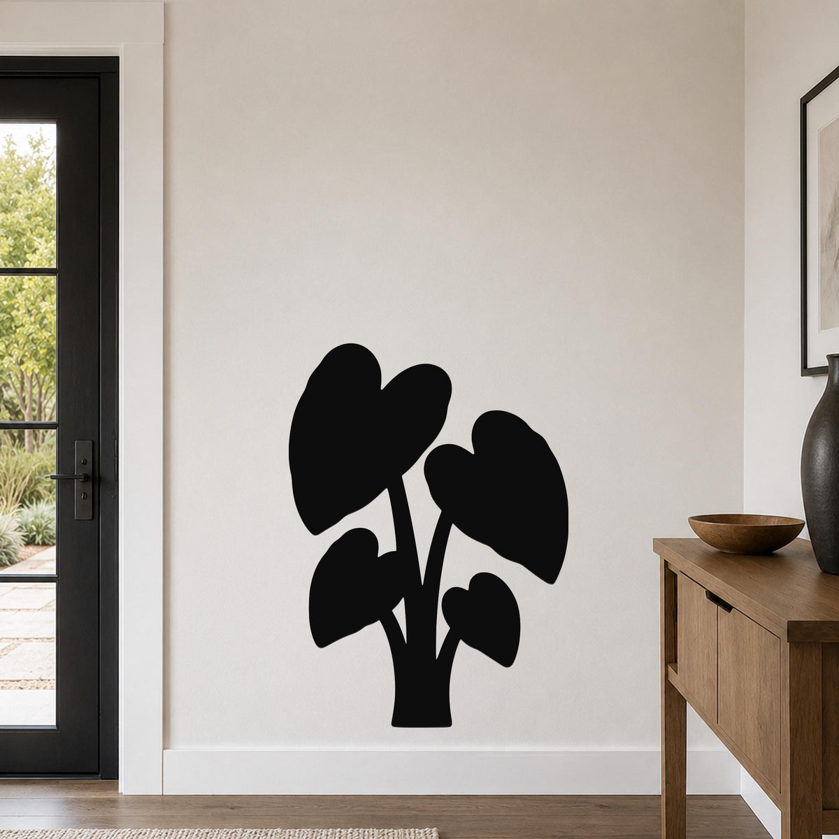 Black metal wall art in the silhouette of an elephant ear plant with large heart-shaped leaves, mounted on a living room wall above a wooden console table.