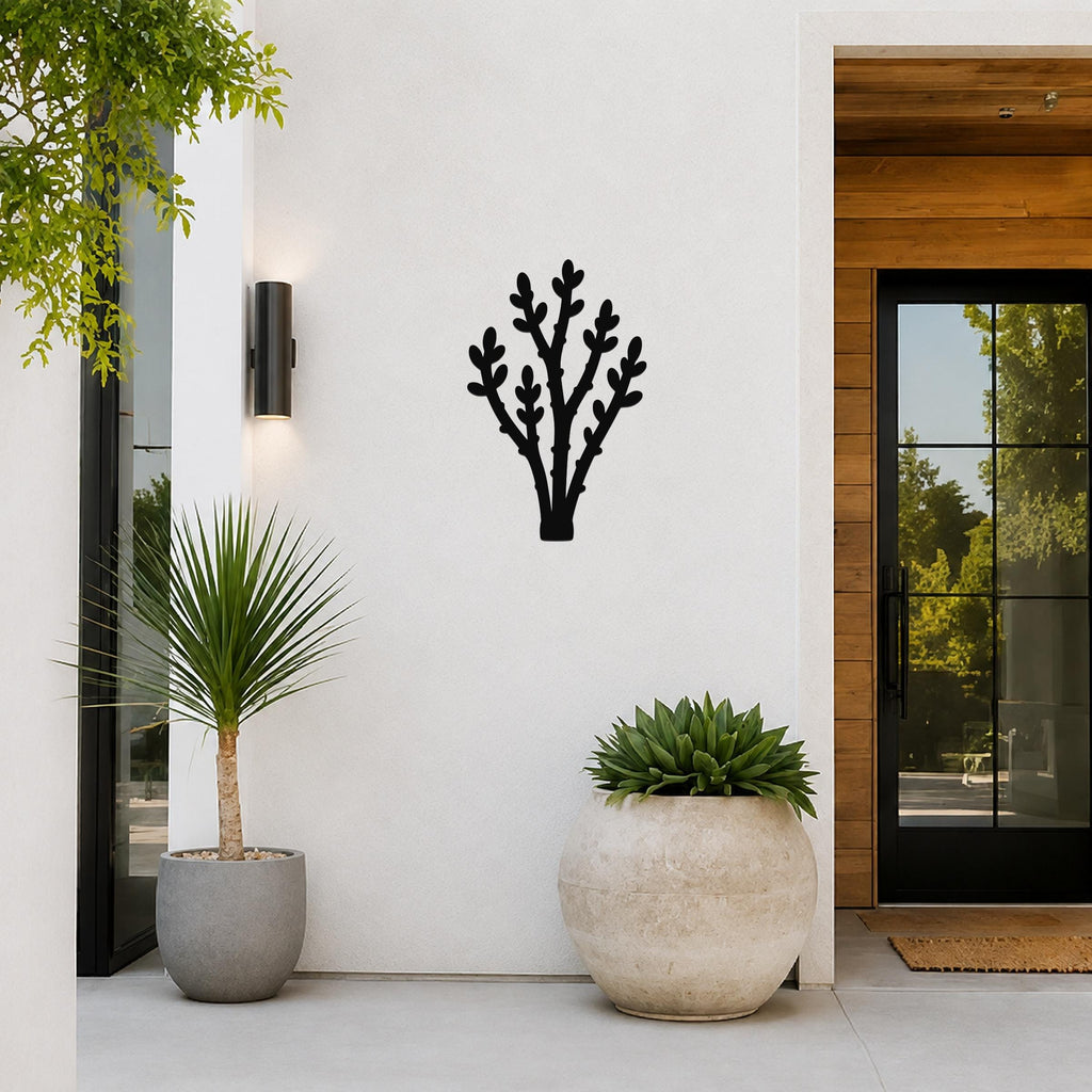 Black metal wall art in a desert branch silhouette, mounted on a white exterior wall above two potted plants near a glass door.