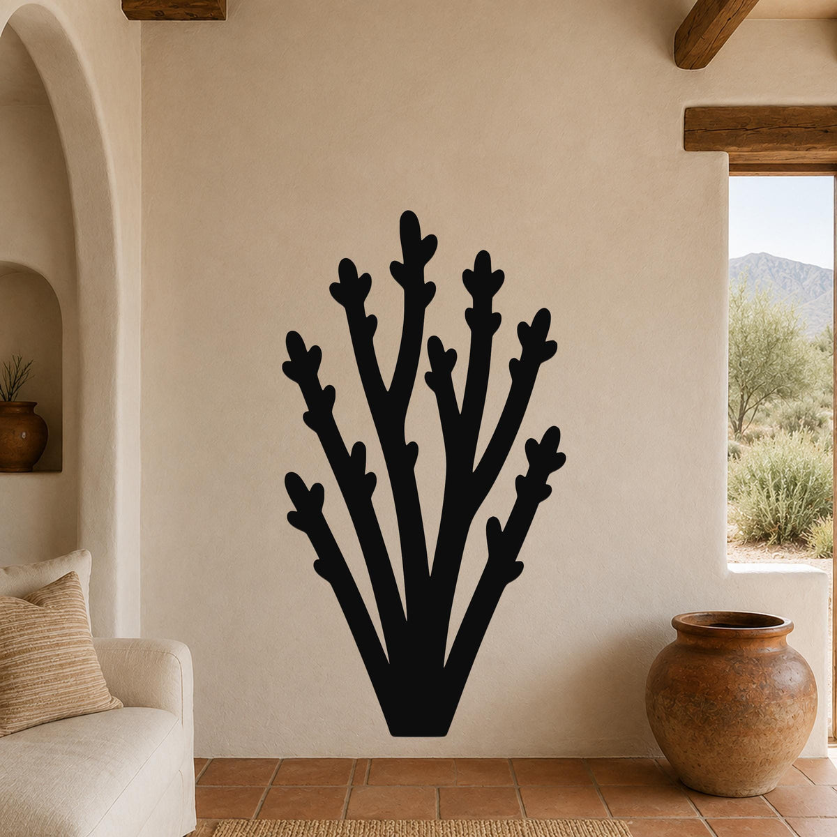 Black metal wall art in a desert branch silhouette, mounted on a beige stucco wall in a living room with terracotta flooring and wooden beams.