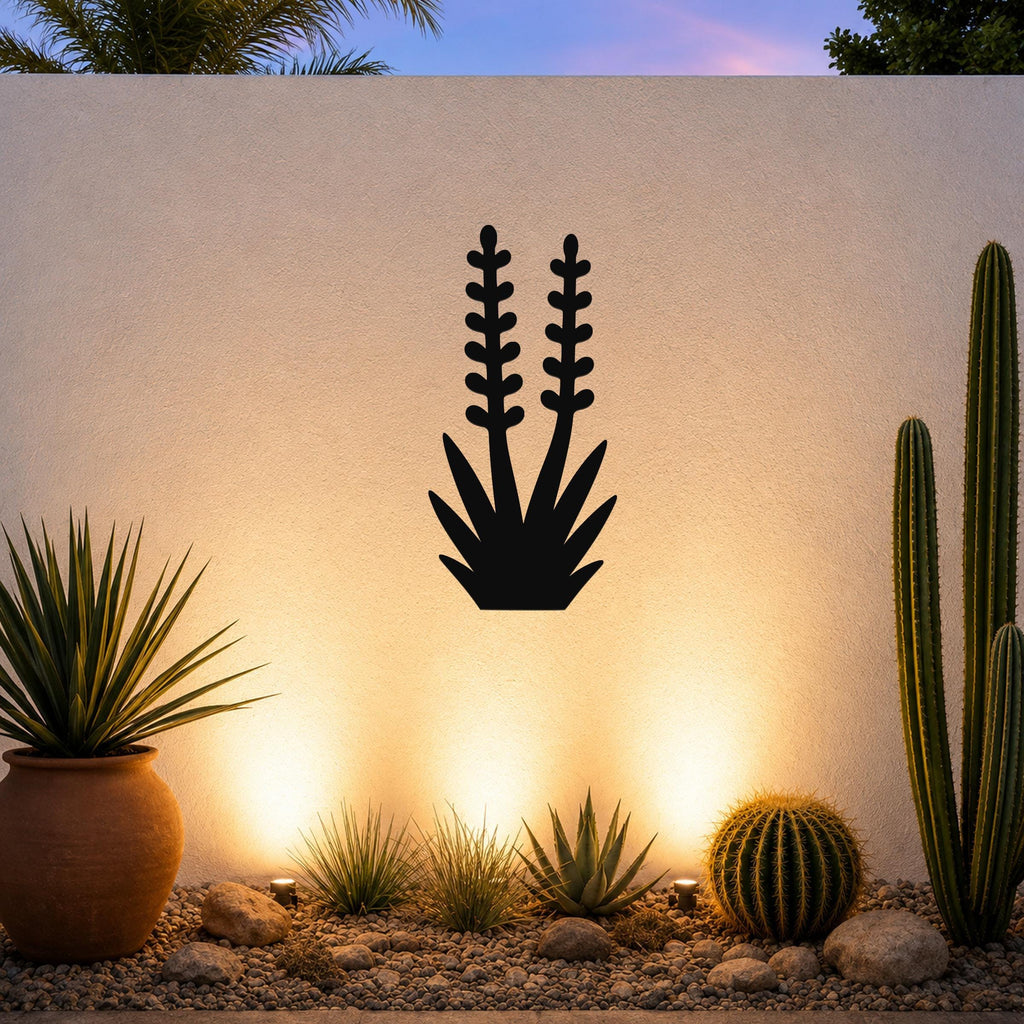 Black metal wall art in the silhouette of a blooming yucca plant with two tall flower stalks, mounted on a textured outdoor wall above a desert garden with cacti and warm uplighting.