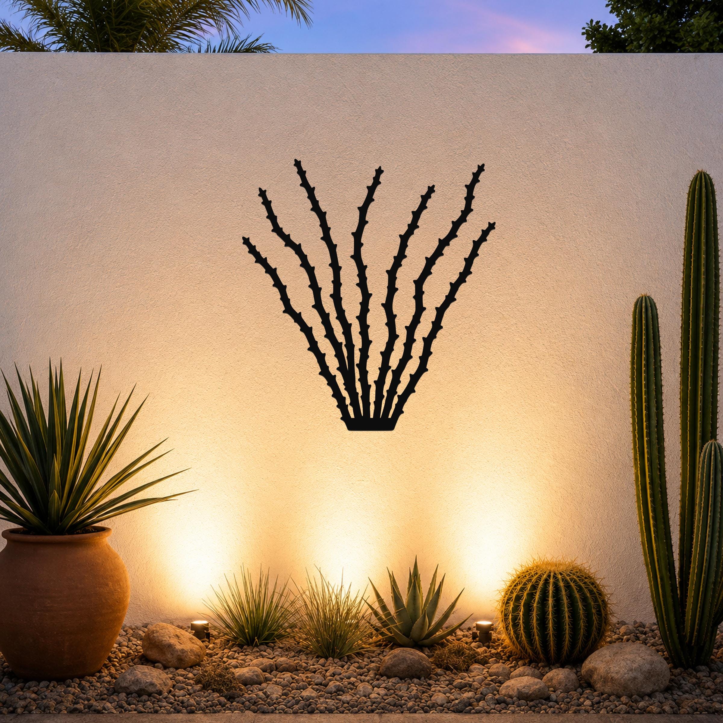 Black metal ocotillo wall art silhouette with spiky branches, mounted on a textured outdoor wall above a desert garden with cacti and warm uplighting.