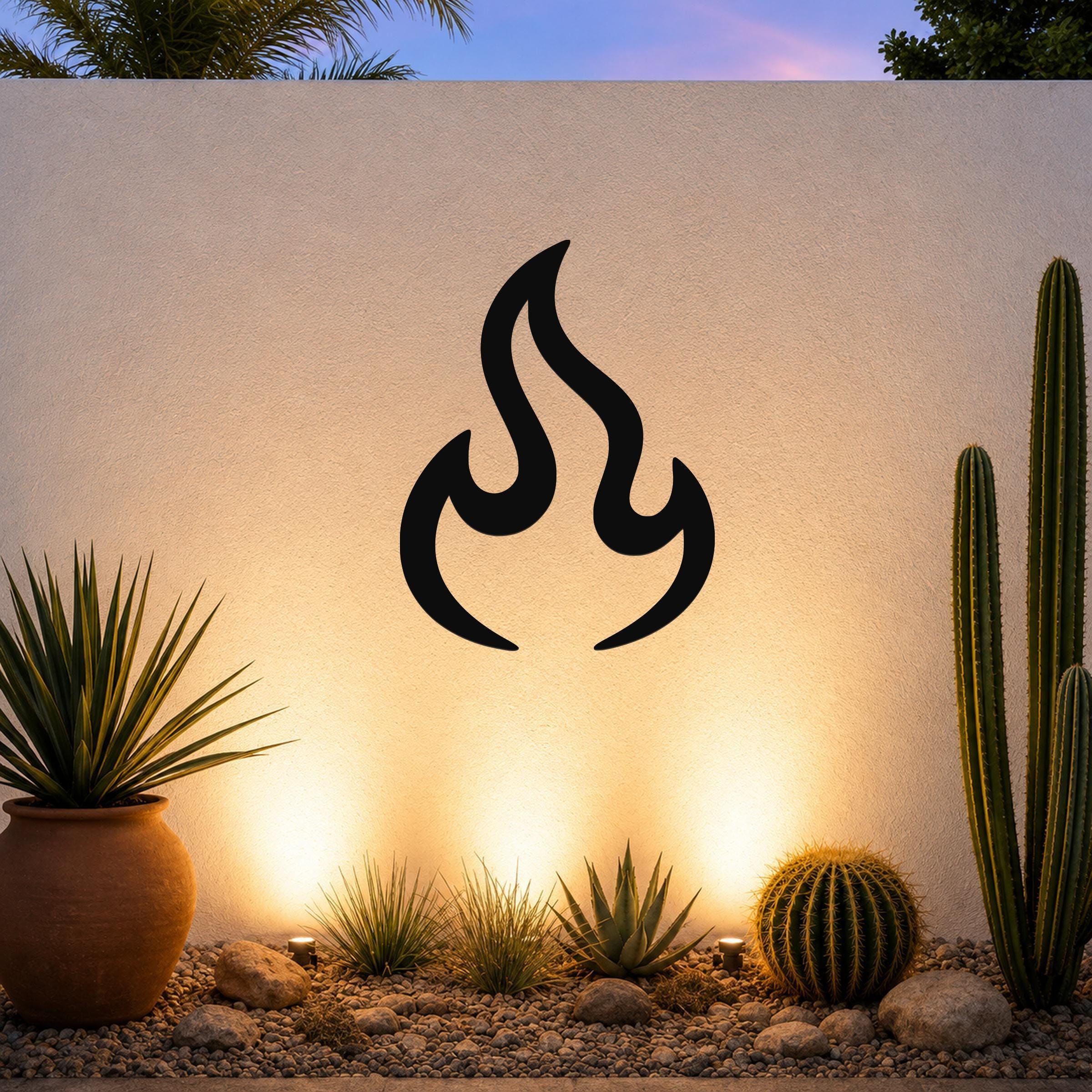 Black metal flame wall art with a bold silhouette design, mounted on an outdoor wall above a desert garden with cacti and warm uplighting.