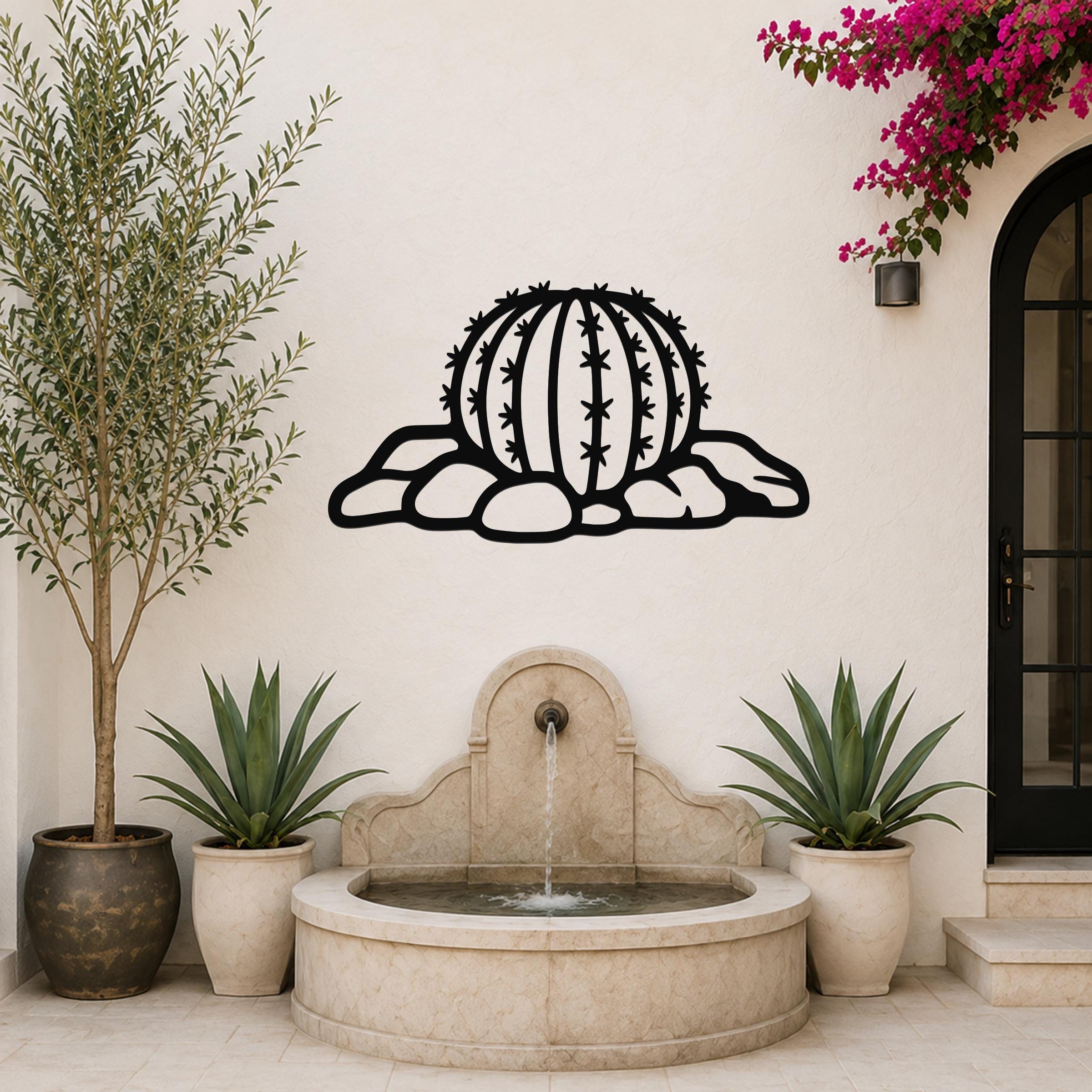 Black metal barrel cactus wall art with spines and rocks cutout, mounted on a white outdoor wall above a stone fountain.