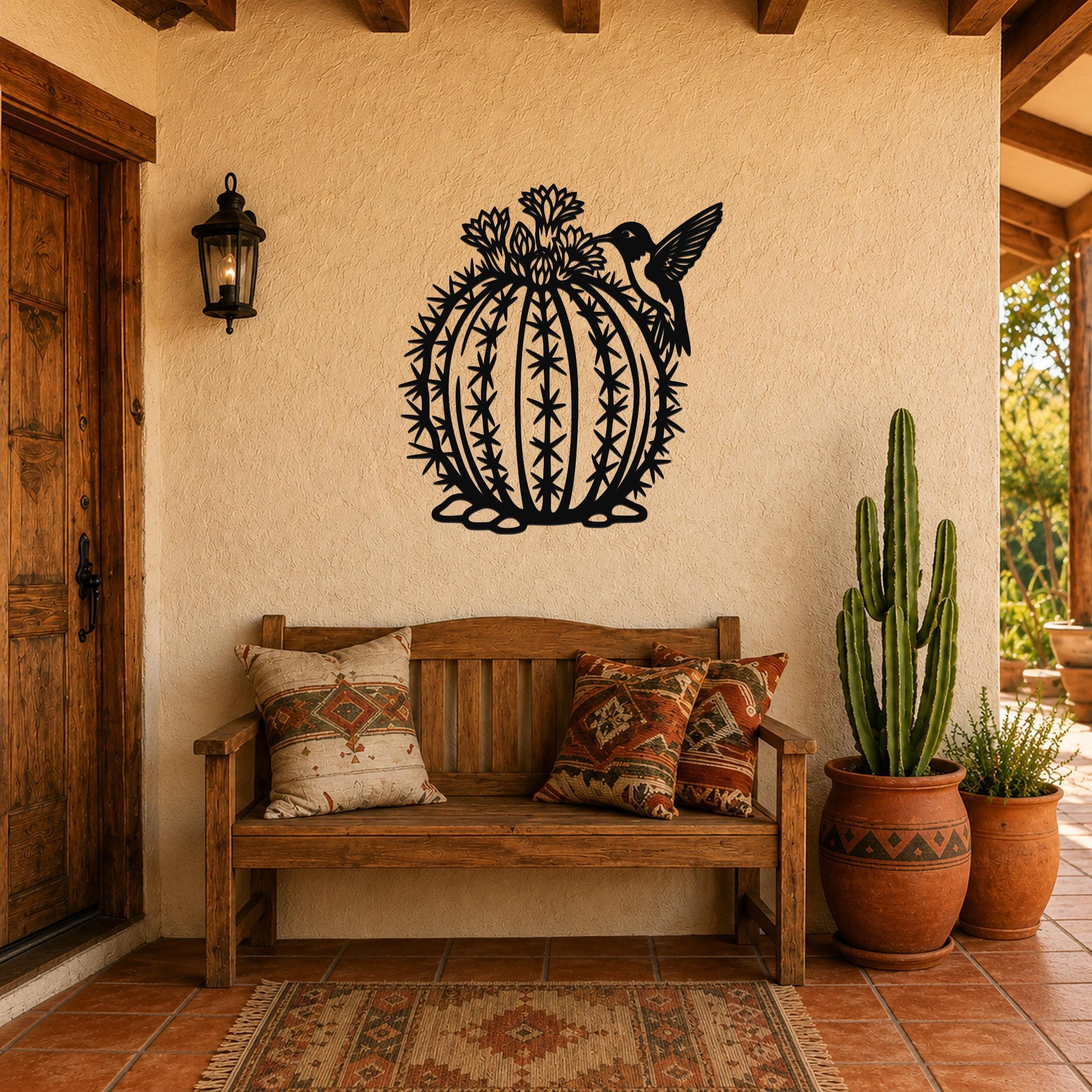 Black metal barrel cactus wall art with hummingbird and flower cutouts, mounted on a stucco wall above a wooden bench in a patio entryway.