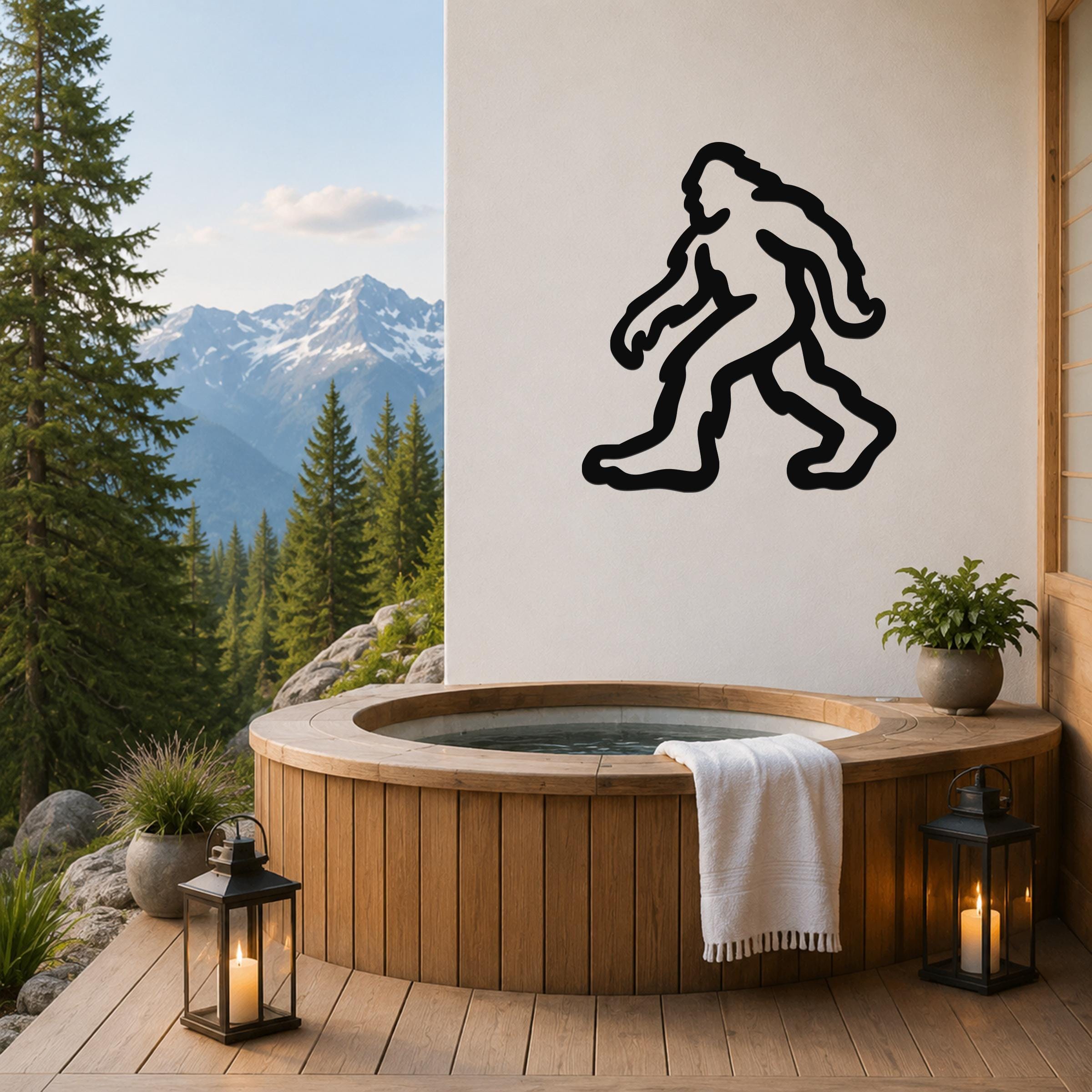 Black metal Bigfoot silhouette wall art mounted on a white wall above a wooden hot tub on a mountain-view deck.