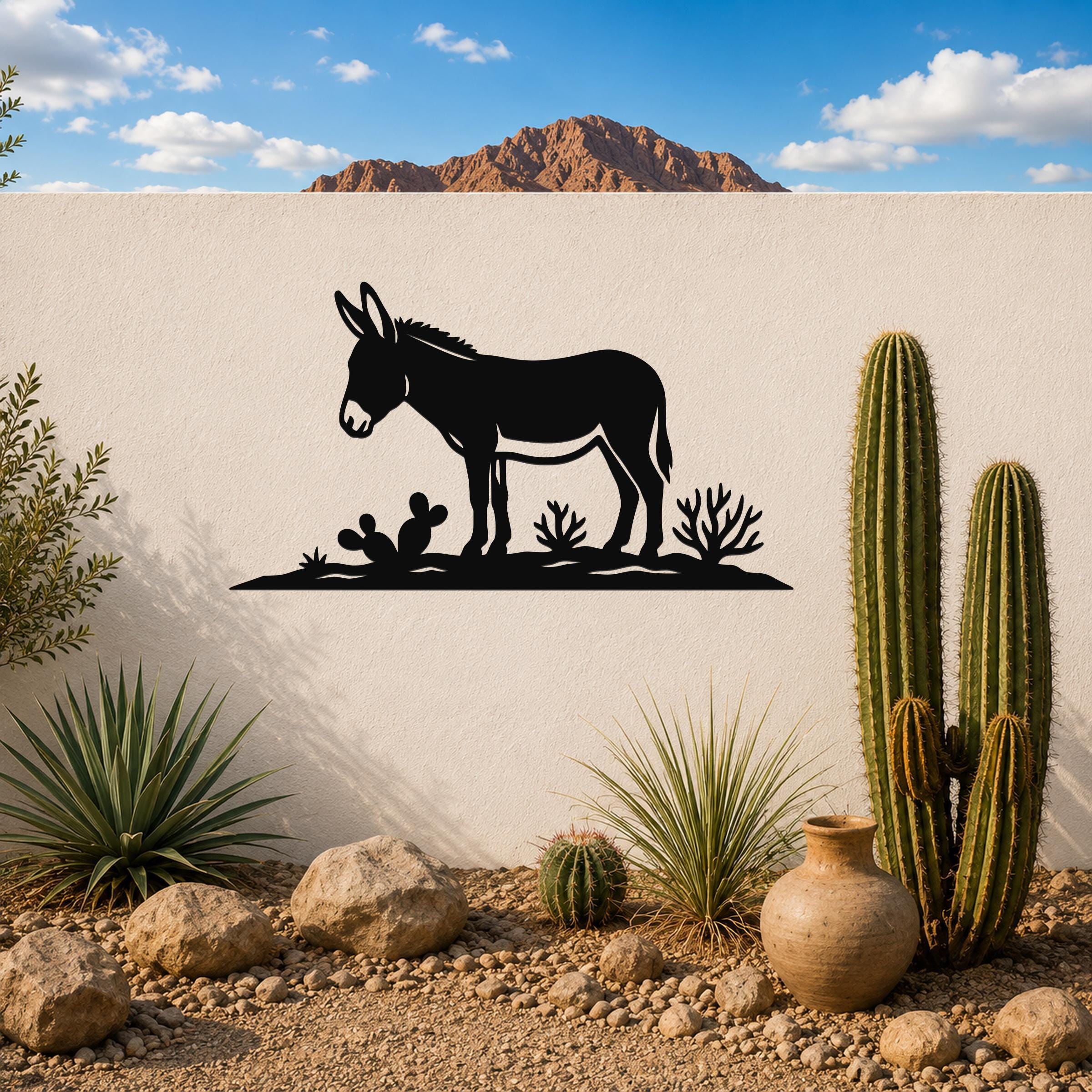 Black metal wall art silhouette of a burro with cacti, mounted on a white outdoor wall in a desert garden setting.