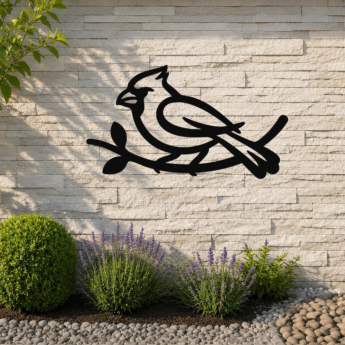 Black metal cardinal wall art in silhouette, perched on a branch with leaf cutouts, mounted on a light stone outdoor wall above a garden bed with lavender and pebbles.