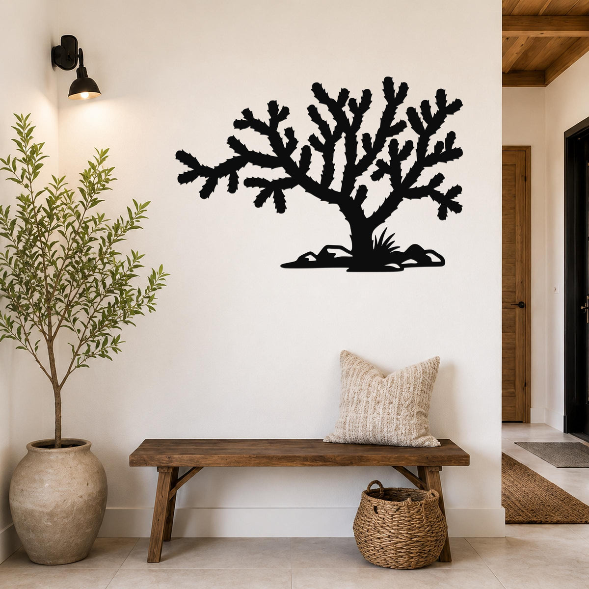 Black metal Cholla cactus wall art silhouette with branching arms and base cutouts, mounted on a white wall above a wooden bench in a hallway.