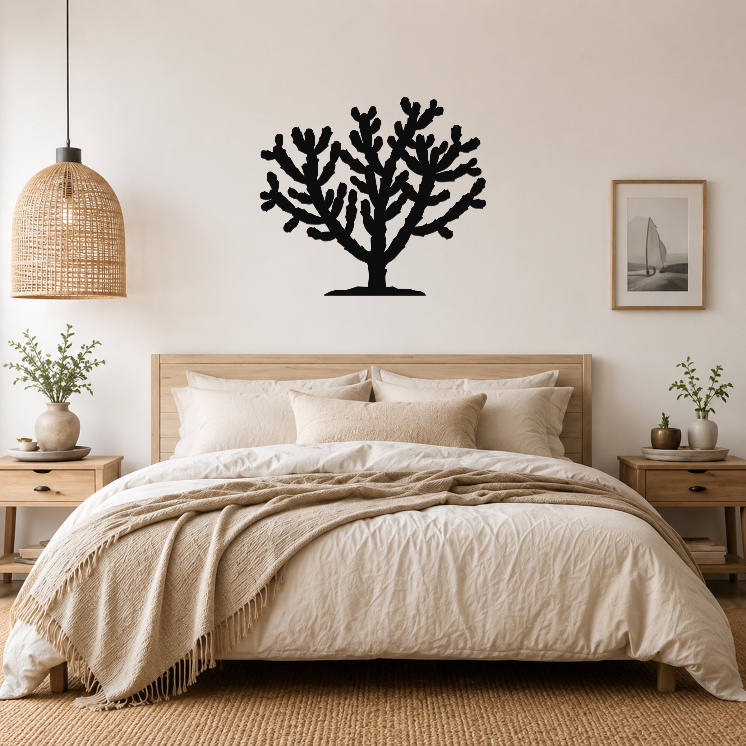 Black metal Cholla cactus wall art silhouette with branching arms, mounted above a wooden bed in a neutral bedroom.