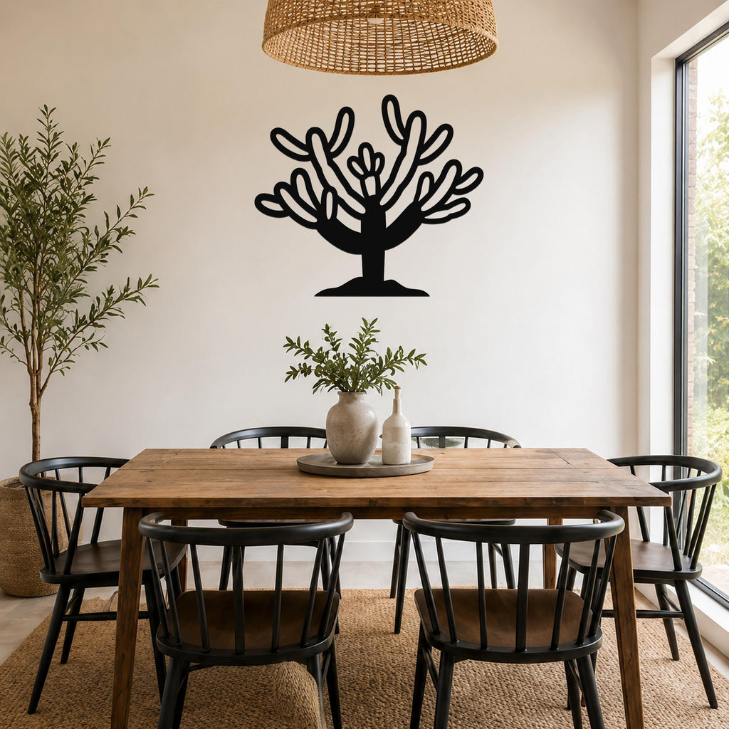 Black metal Cholla cactus wall art with branching cutouts, mounted on a white wall above a wooden dining table in a bright room.