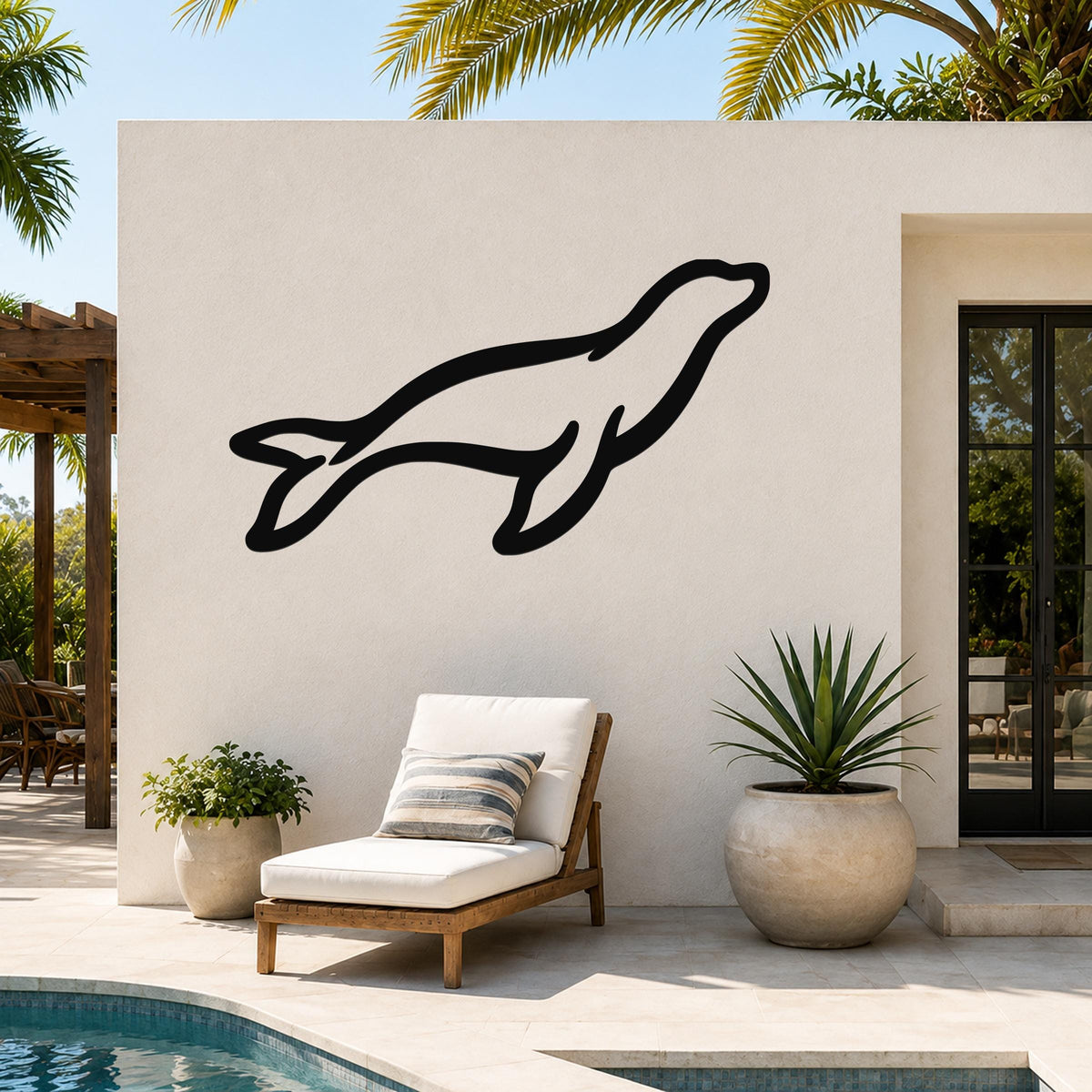 Black metal sea lion wall art silhouette mounted on an outdoor patio wall above a lounge chair, with palm trees and a pool in the background.