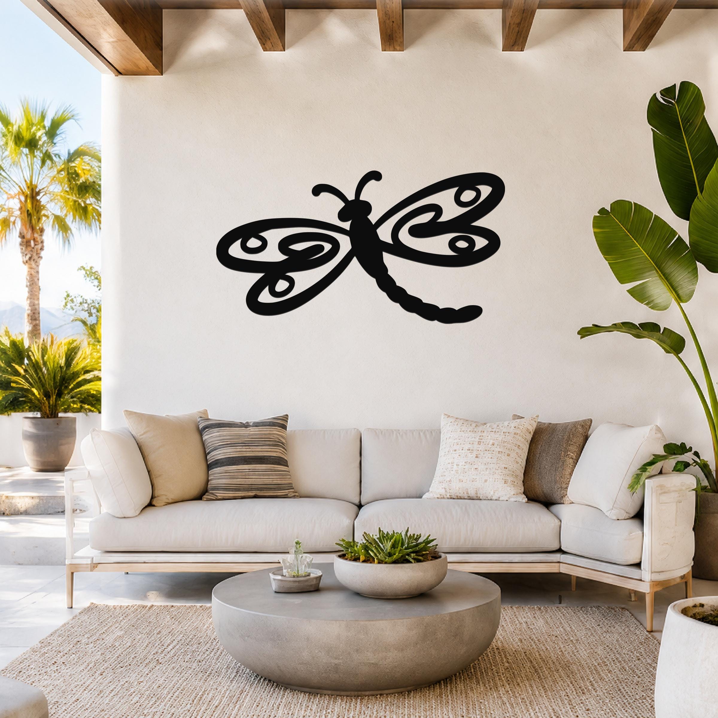 Black metal dragonfly wall art with intricate wing cutouts, mounted on a white wall above a light-colored sofa in a bright living room.