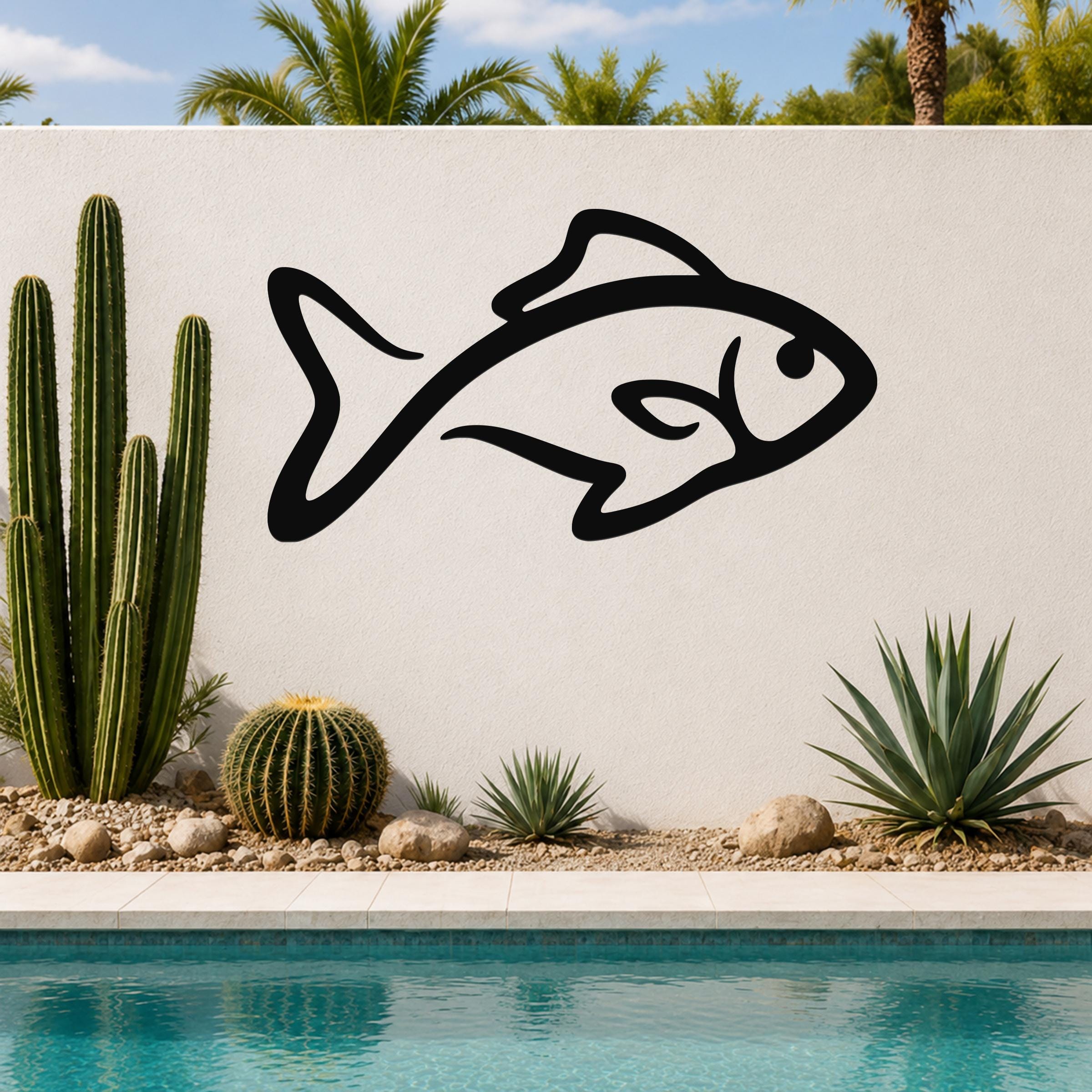 Black metal fish silhouette wall art with internal cutout details, mounted on a white outdoor wall beside a pool and cacti.