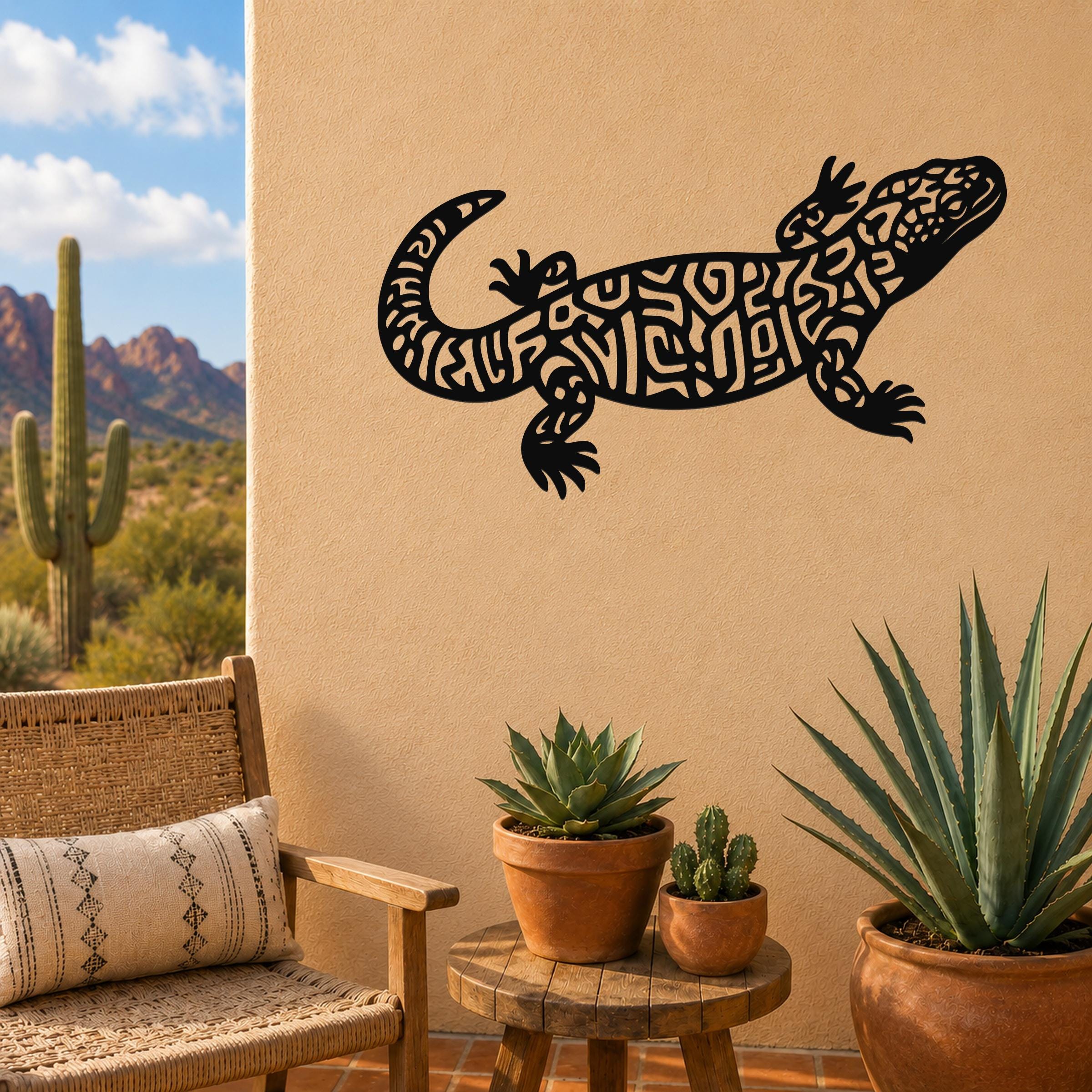 Black metal Gila monster wall art with intricate geometric cutouts, mounted on a tan outdoor wall above a small wooden table with potted succulents.