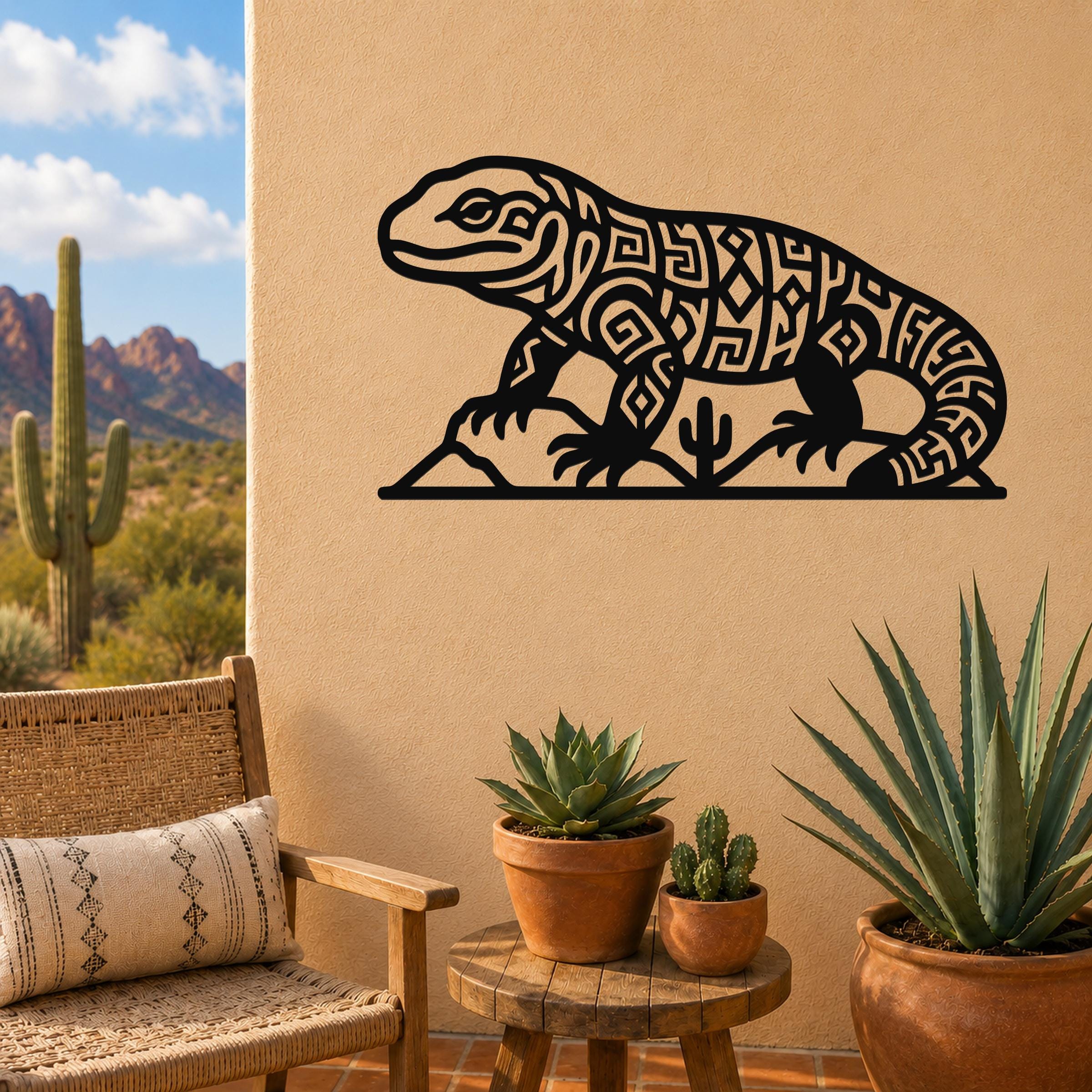 Black metal wall art silhouette of a Gila monster with intricate geometric cutouts, mounted on a tan outdoor wall above a small table with potted succulents and a desert landscape view.