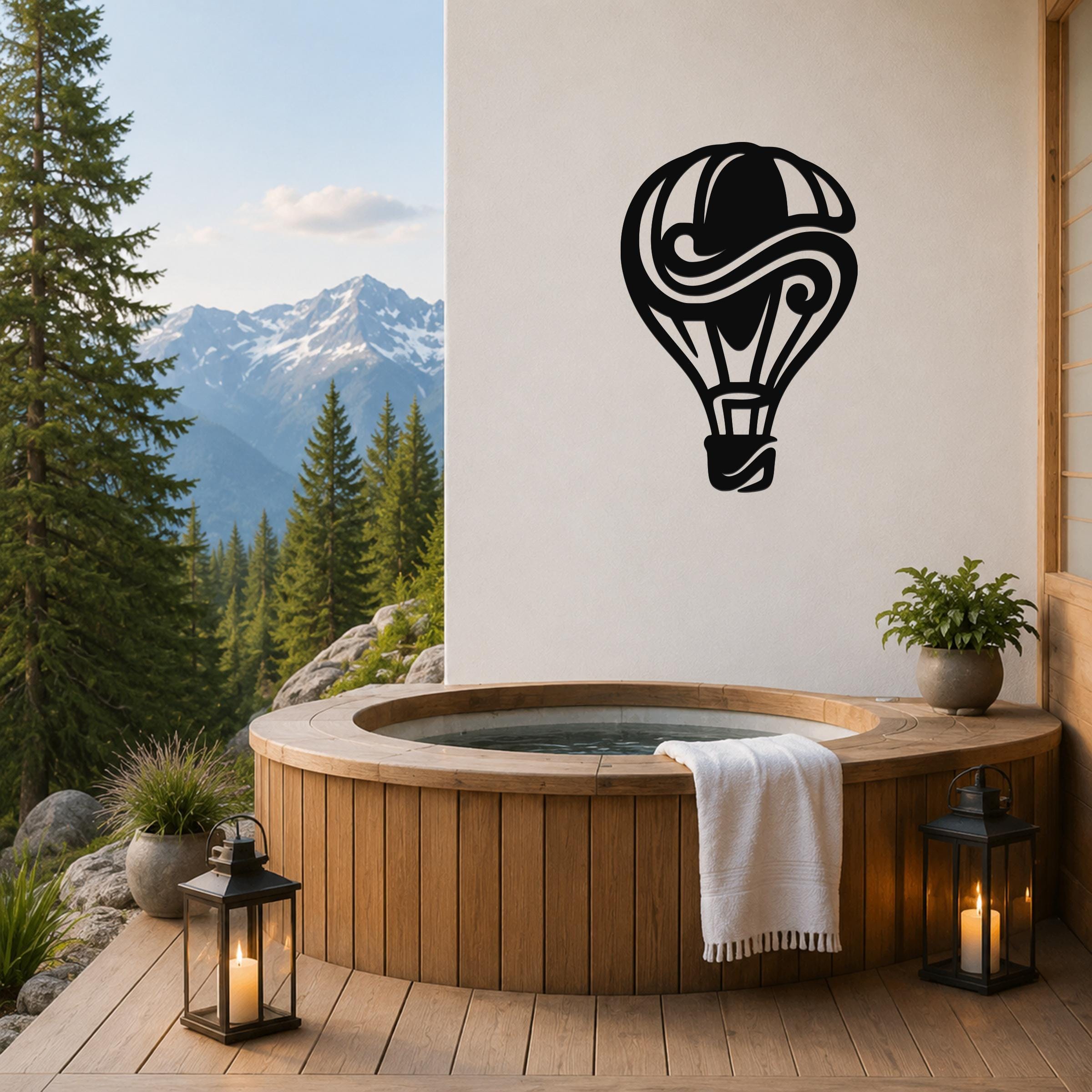 Black metal hot air balloon wall art with swirling internal cutouts, mounted on a white wall above a wooden hot tub on a mountain-view deck.
