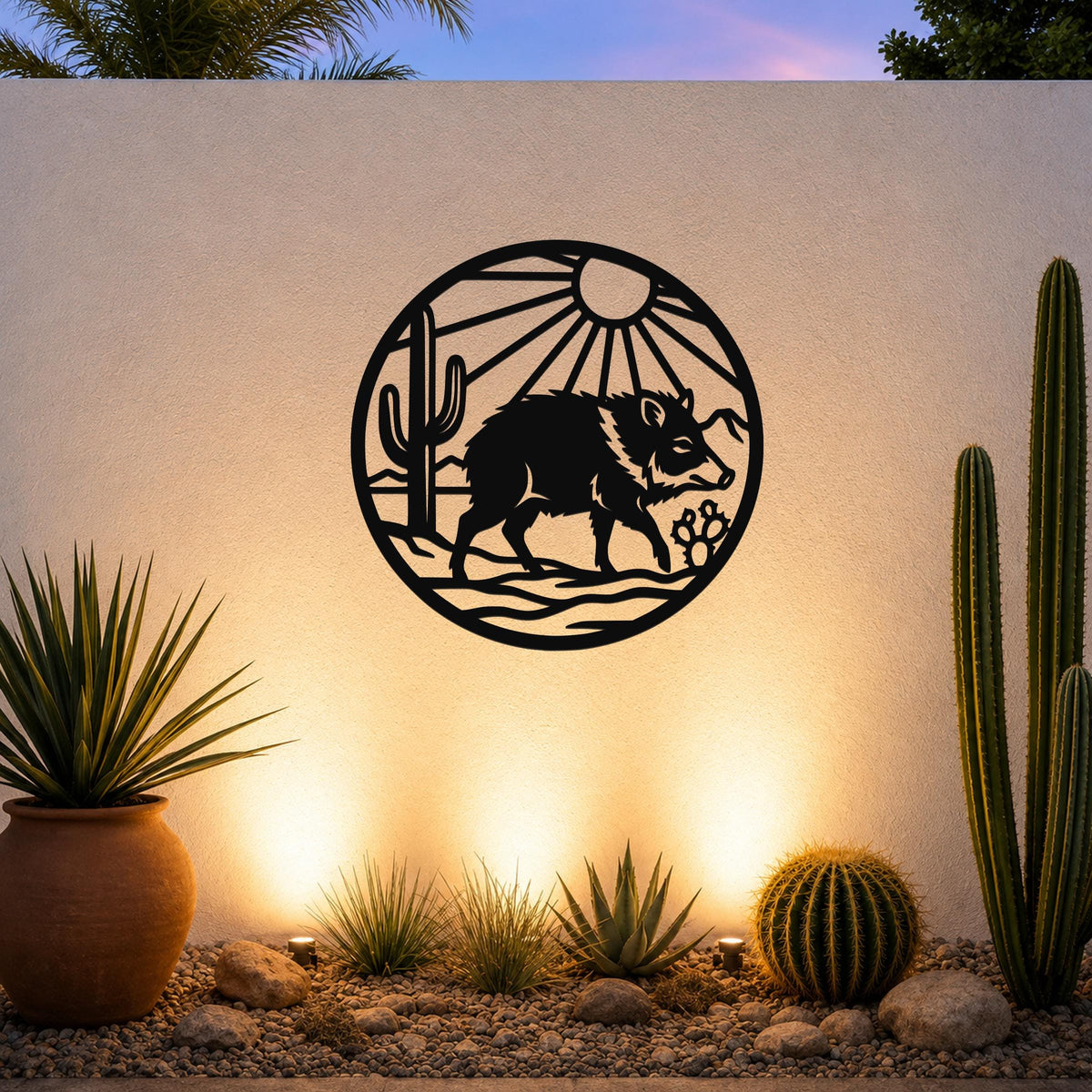 Black metal circular wall art featuring a javelina silhouette, cactus, and sunburst rays, mounted on an outdoor wall above a desert garden with cacti and warm uplighting.