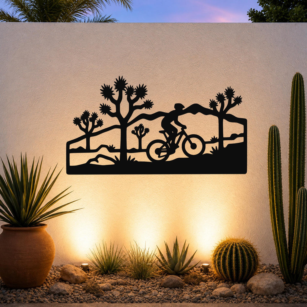 Black metal wall art with a silhouette of a cyclist and Joshua trees, mounted on an outdoor wall above a desert garden with cacti and warm uplighting.