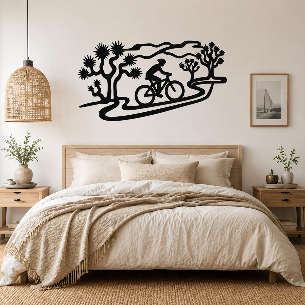 Black metal wall art silhouette of a cyclist on a winding path through Joshua trees, mounted above a bed in a bedroom.