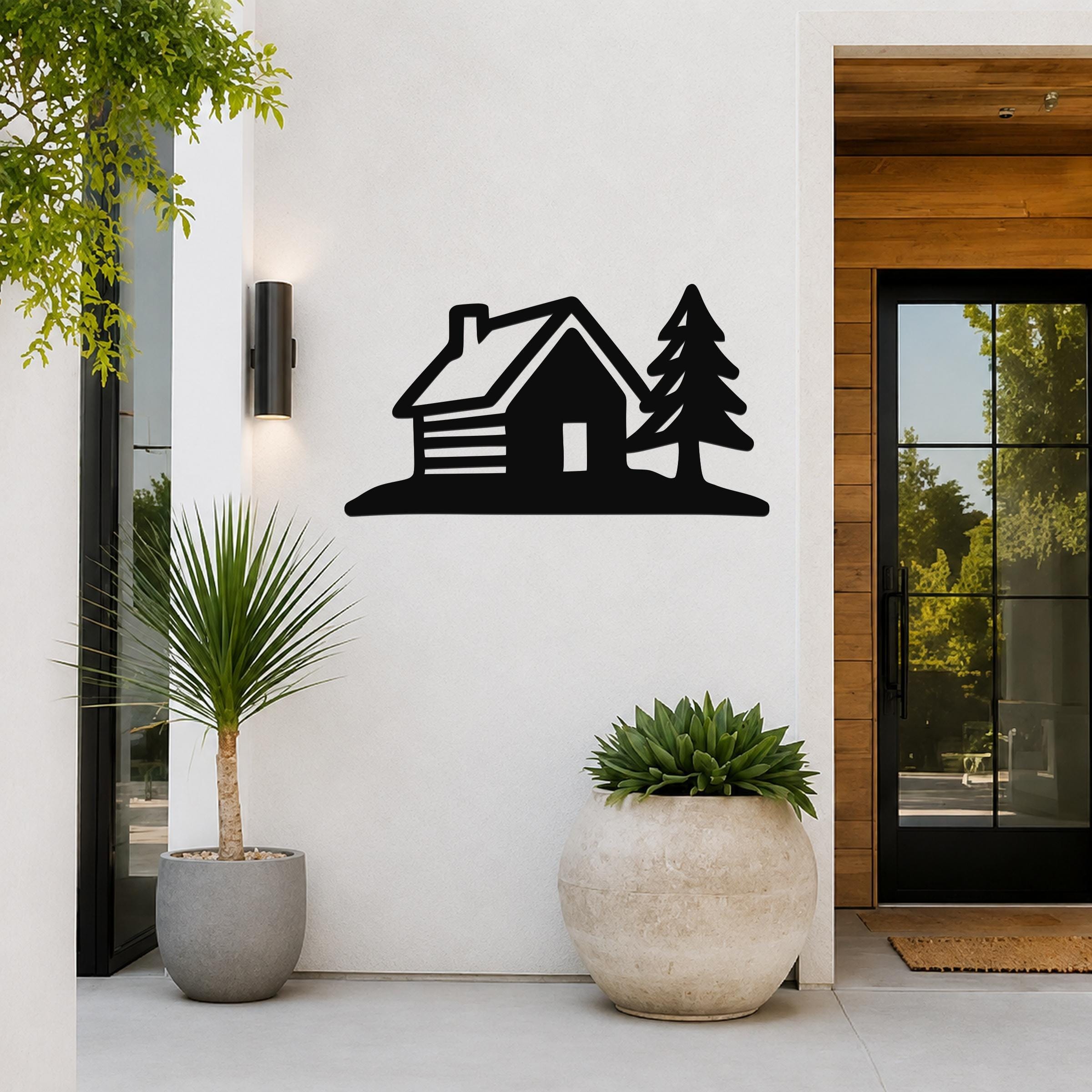 Black metal wall art silhouette of a cabin and pine tree, mounted on a white exterior wall beside a glass door and potted plants.