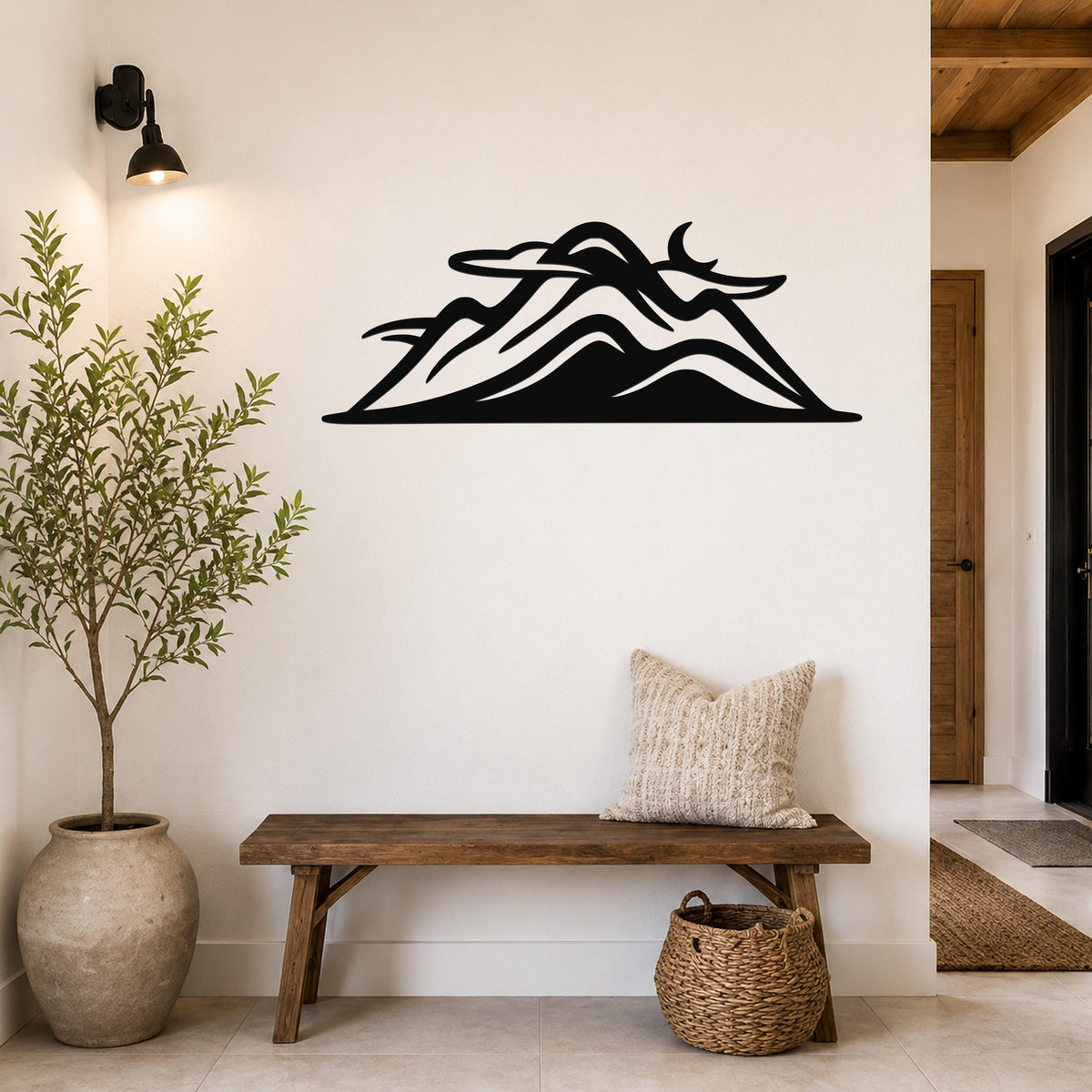 Black metal mountain silhouette wall art with layered peaks and cutout details, mounted on a white wall above a wooden bench in a rustic entryway.