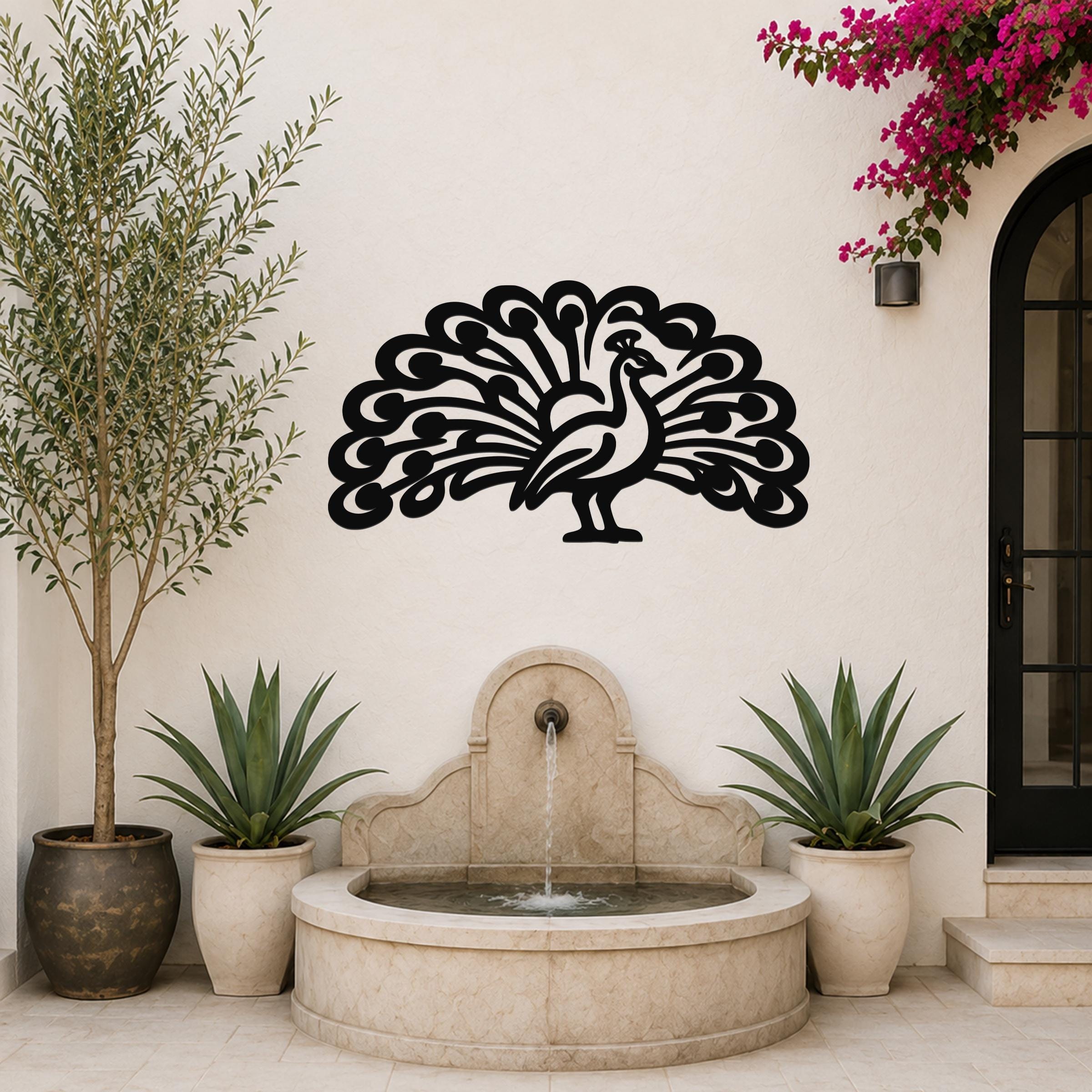 Black metal peacock wall art with intricate feather cutouts, mounted on a white outdoor wall above a stone fountain.