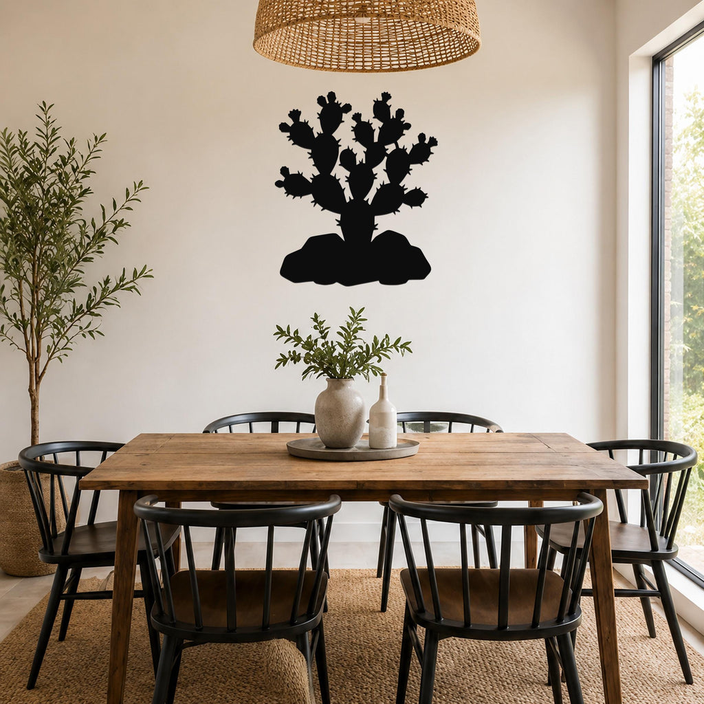 Black metal prickly pear cactus wall art silhouette with spines and pads, mounted on a white wall above a wooden dining table in a bright dining room.