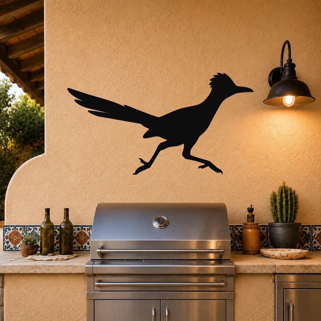 Black metal roadrunner wall art silhouette mounted on a stucco outdoor wall above a stainless steel grill, with a lit wall sconce and cactus nearby.