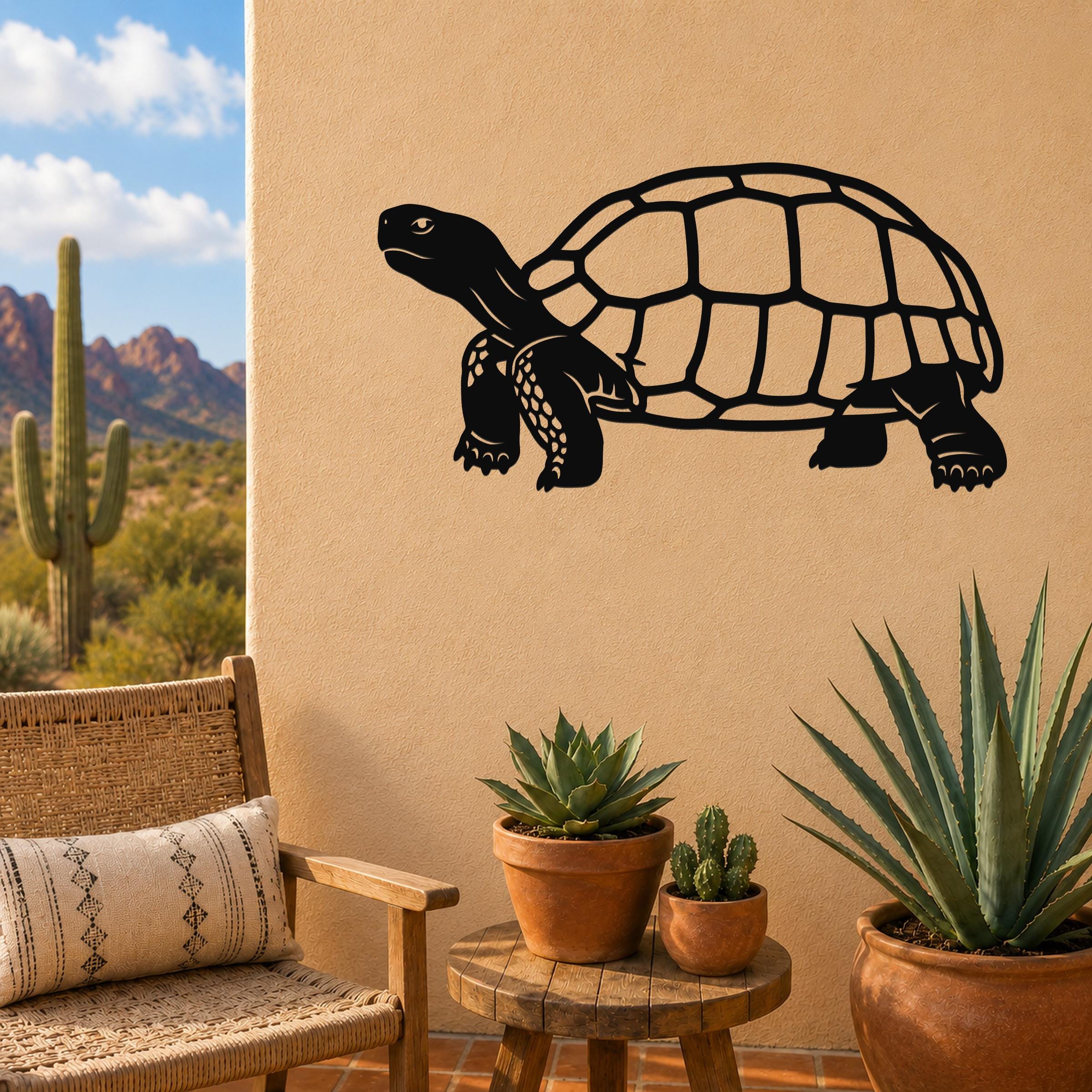 Black metal tortoise wall art with detailed shell and limb cutouts, mounted on a beige outdoor wall above a small wooden table with potted succulents.