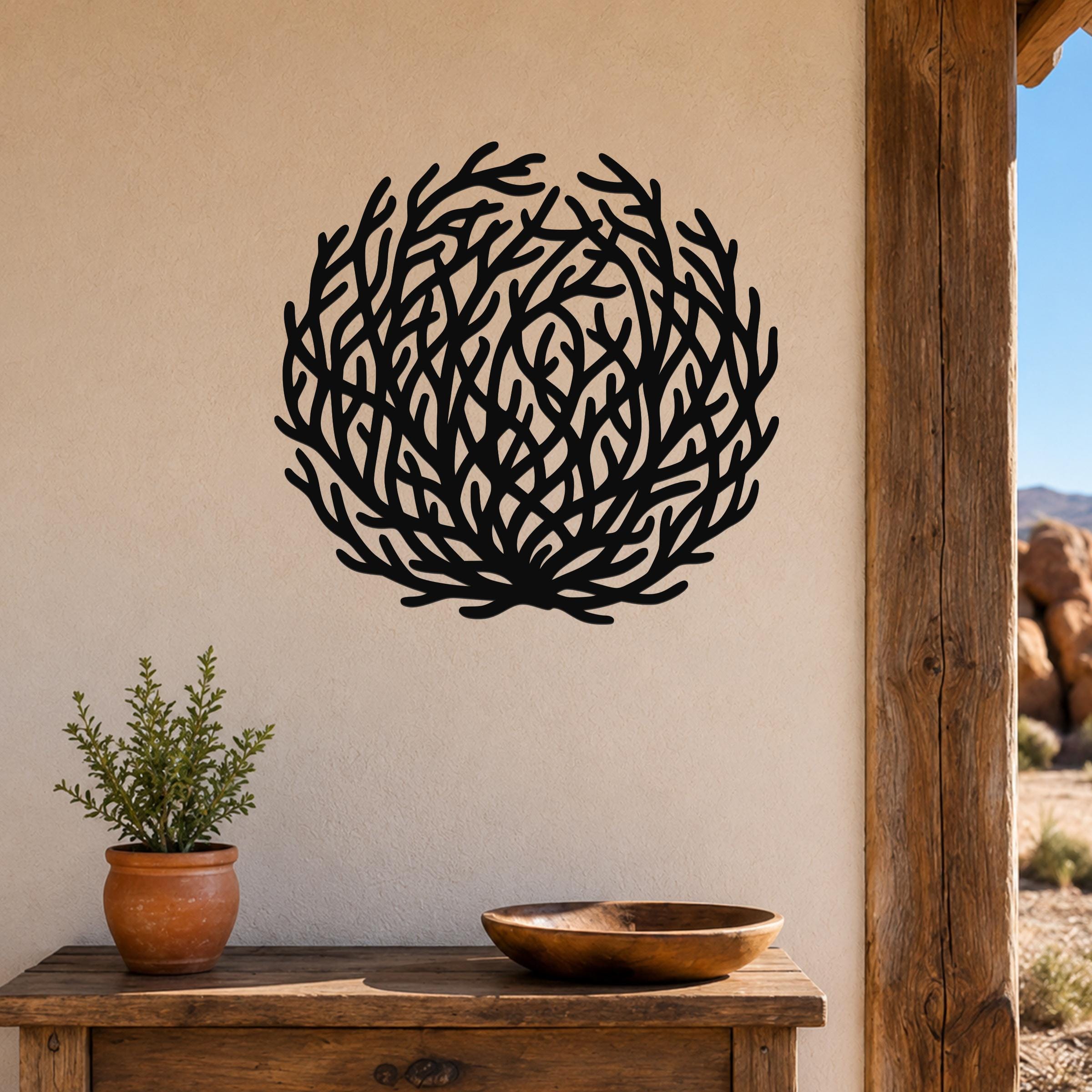 Black metal wall art in a circular tumbleweed silhouette with branching cutouts, mounted on a beige wall above a wooden table with a potted plant and bowl.