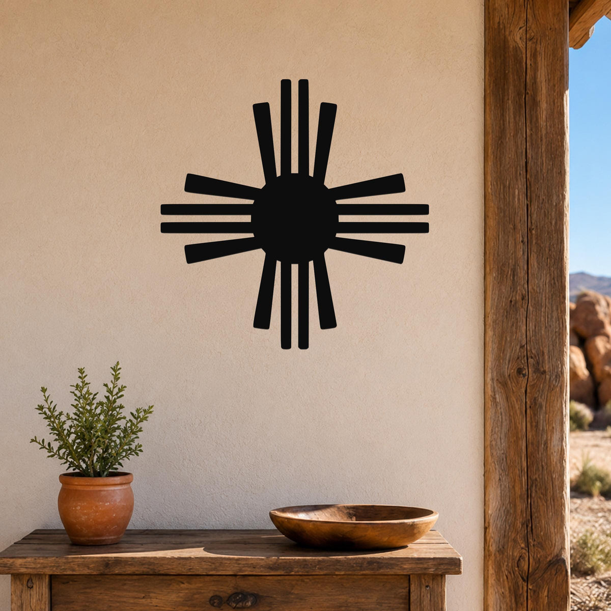 Black metal Zia Sun symbol wall art with eight rays, mounted on a beige stucco wall above a wooden table with a potted plant and bowl.